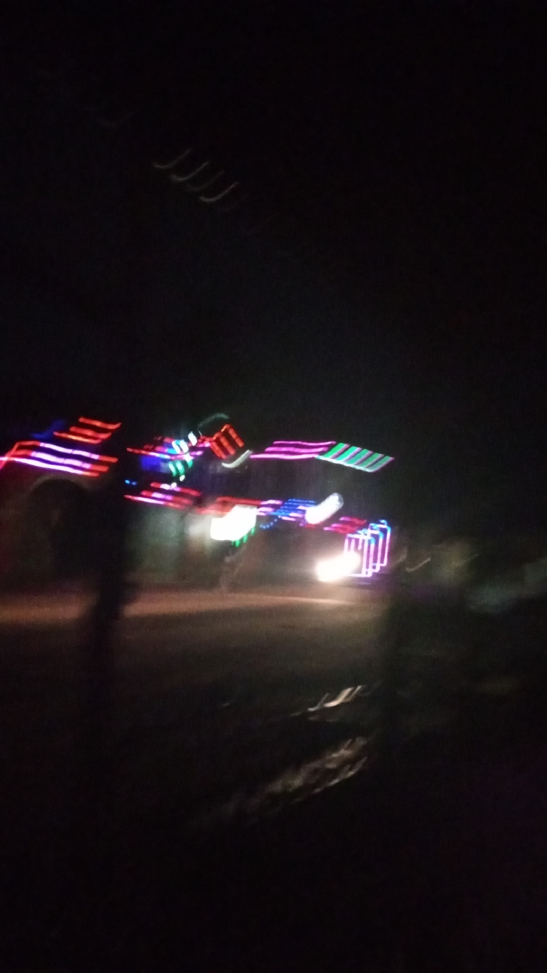This is a highly blurred nighttime photograph, likely taken in Narayi, Nigeria, capturing a scene dominated by vibrant streaks of colorful lights against a dark background. The significant motion blur suggests either a fast-moving subject or the camera itself was in motion during a long exposure.
The most prominent subjects are vivid, elongated streaks of red, pink, green, blue, and white lights that cut across the mid-ground. These lights appear to be decorative illuminations on a structure or a vehicle, creating a dynamic and festive visual. In the lower center-right, a strong white light is visible, consistent with a vehicle's headlight. A faint, dark, human-like silhouette, heavily blurred, can be discerned on the left side of the frame. The ground or road surface is dimly visible in the lower portion of the image. In the upper left quadrant, some subtle, curved light trails can be seen, possibly reflections or distant lights.
The scene takes place outdoors at night, indicating a festive or celebratory event. The colorful lighting strongly suggests a parade, procession, or a decorated vehicle moving through a street or open area. The overall impression is one of movement and vibrancy associated with a nighttime celebration. There is no discernible text within the image.