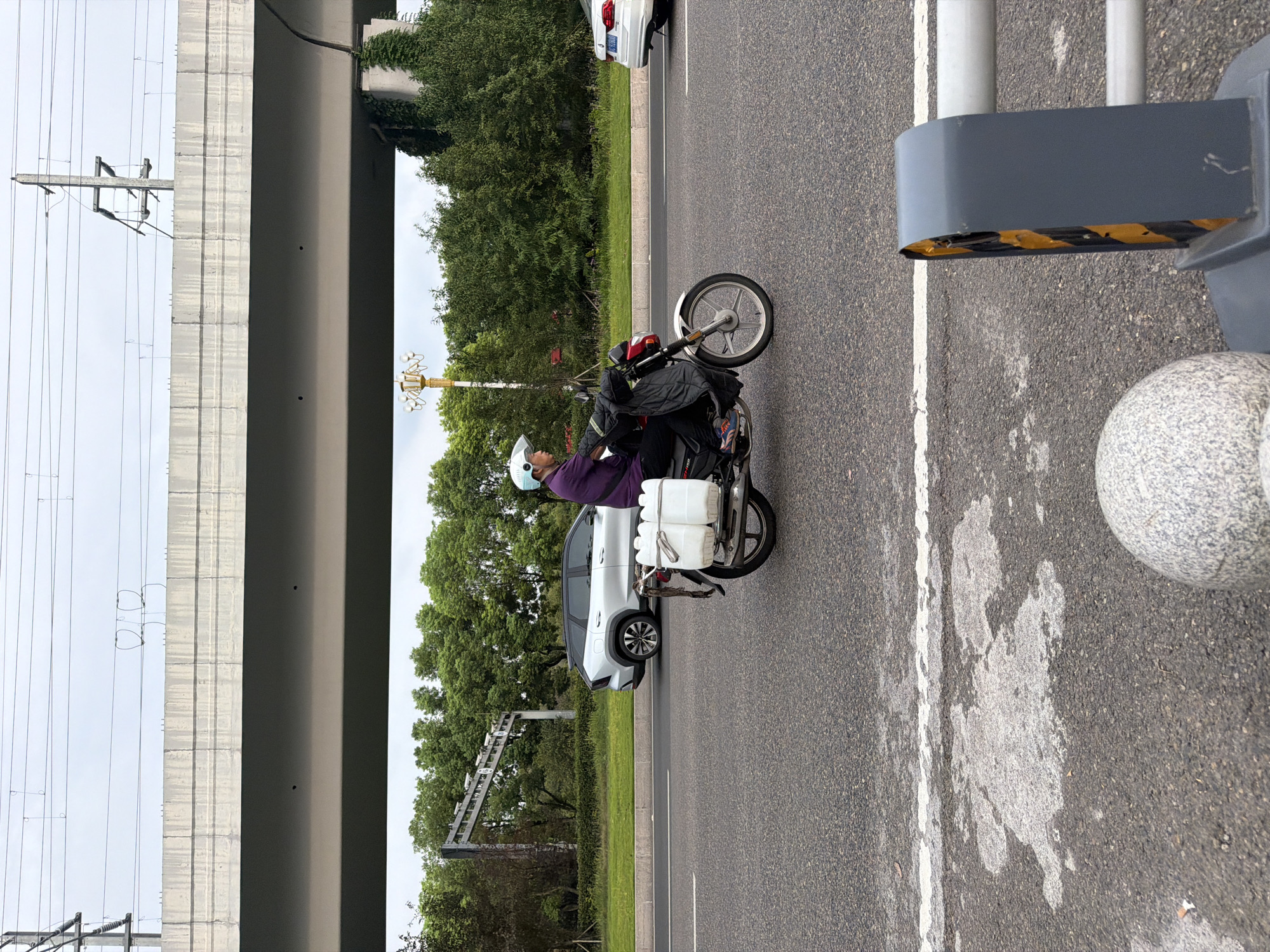 A person wearing a light blue helmet and a purple jacket rides a motorcycle on a paved road in Jiangjin, China. The motorcycle carries two large white plastic containers strapped to its rear. The road is lined with trees and a highway overpass is visible in the background. A silver SUV is parked on the side of the road. The sky is overcast.