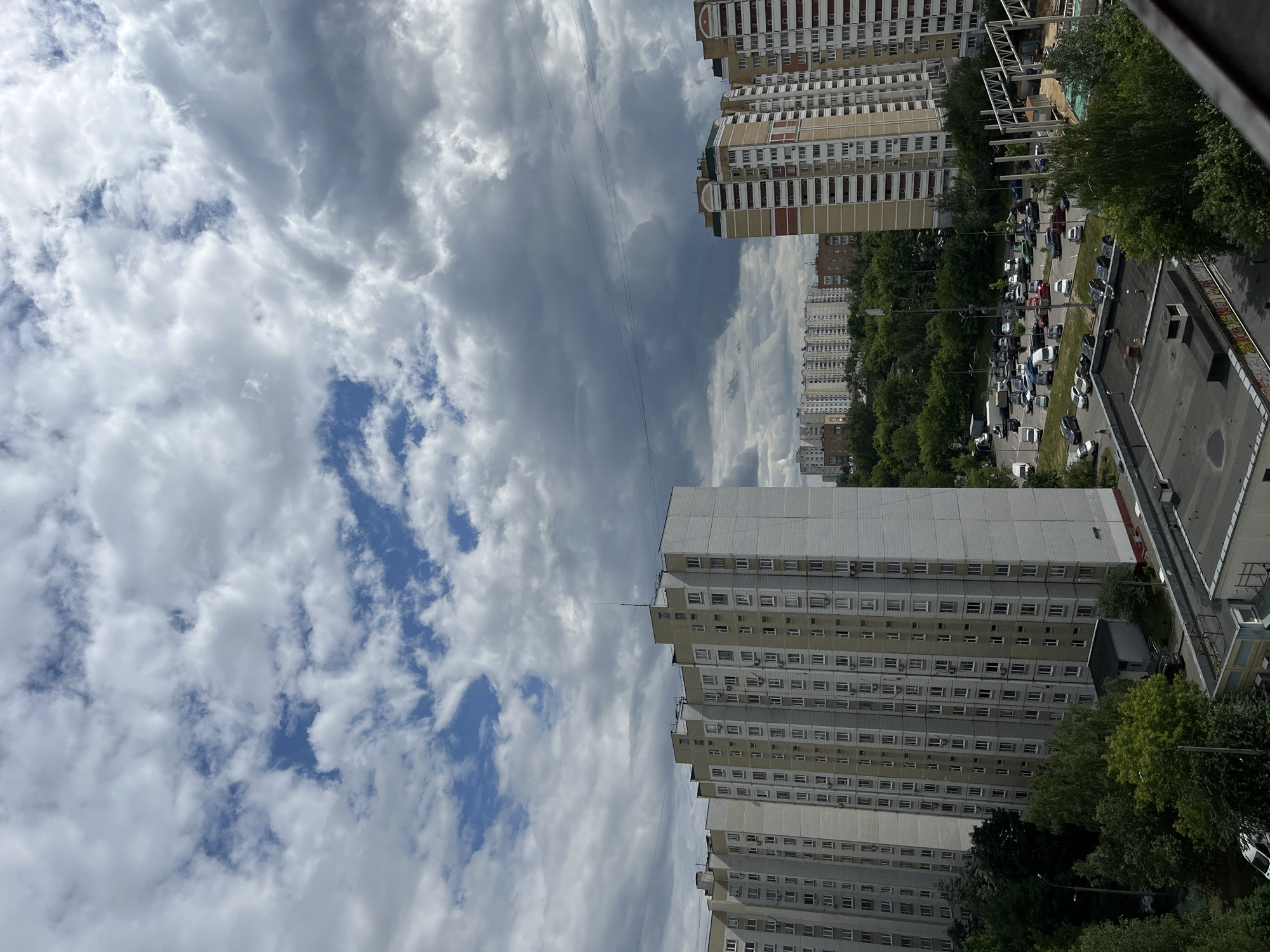 The image captures a high-angle view of a residential area in Moscow, Russia. In the foreground, several multi-story apartment buildings dominate the scene. The building on the left is a tall, pale yellow structure with many uniformly spaced windows. To its right, another similar building, slightly shorter, displays a similar architectural style with light gray and white facades. Further to the right, a more colorful apartment building stands out with its beige and red striped pattern.
Below the buildings, a dense canopy of green trees stretches across the middle ground, interspersed with glimpses of roads and parked cars. A parking lot filled with various vehicles, including cars and vans, is visible in front of the buildings. In the far distance, a panorama of more apartment buildings can be seen, characteristic of Moscow's urban landscape. The sky above is filled with dramatic, heavy clouds, with patches of blue sky visible through them, suggesting overcast or potentially stormy weather. Power lines crisscross the sky between the buildings. The overall impression is that of a densely populated urban environment on a cloudy day.