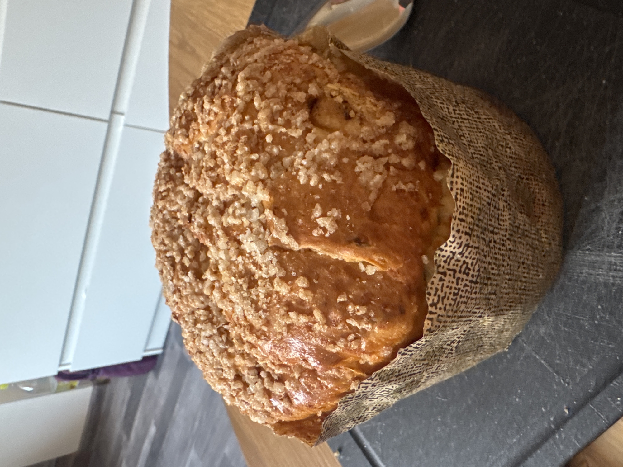 A close-up, indoor shot presents a sweet bread loaf in a paper wrapper, speckled with sugar crystals on its crust, placed on a dark gray surface. The background reveals a partially visible white-tiled wall and a wooden surface. The bread is round with a golden-brown hue. The location is not discernible from the image, so it is impossible to determine if it is in Varna, Bulgaria.