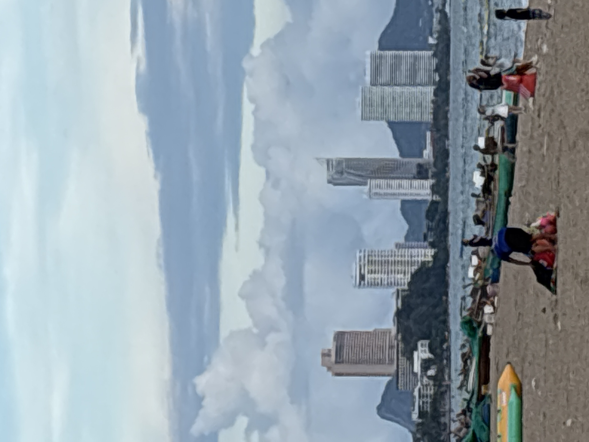 This is a photograph taken at Pattaya Beach in Muang Pattaya, Thailand. The image captures a coastal scene with a sandy beach in the foreground, the sea in the mid-ground, and a city skyline with numerous high-rise buildings and mountains in the background. The sky is partly cloudy, with large, fluffy clouds dominating the upper portion of the frame.
On the beach, several people are visible. In the foreground, a person is kneeling next to some luggage or bags. Further back, near the water's edge, a group of people are standing and walking. Some individuals appear to be interacting with each other. There are also several boats, some of which appear to be fishing boats, pulled up onto the sand. A yellow and green inflatable watercraft is positioned on the left side of the beach.
The city skyline features a variety of modern architecture, including several tall buildings and lower structures. The presence of mountains behind the city suggests a coastal mountainous region. The sea appears calm, with gentle waves lapping at the shore. The overall lighting suggests it may be late afternoon or early evening, given the soft light and the atmospheric conditions. The image has a somewhat soft focus, giving it a slightly painterly quality.