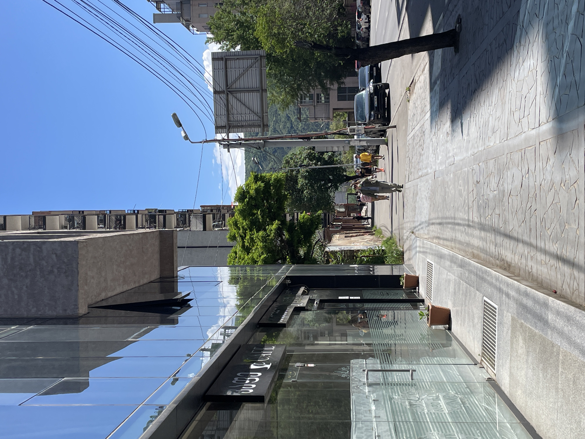 The image depicts a street in Tbilisi, Georgia. The foreground features a modern glass-walled building with signage in Georgian script, possibly a cafe or shop. Reflections of the sky and surrounding structures are visible in the glass. The sidewalk adjacent to the building is paved with light-colored stone tiles. Small potted plants are placed near the base of the building.
Further down the street, pedestrians are walking. Vehicles are parked on the side of the road. A utility pole with overhead wires and a street lamp is visible. Trees line the street, and a billboard structure stands above the street. In the background, a mountain is visible. The sky is clear and blue.