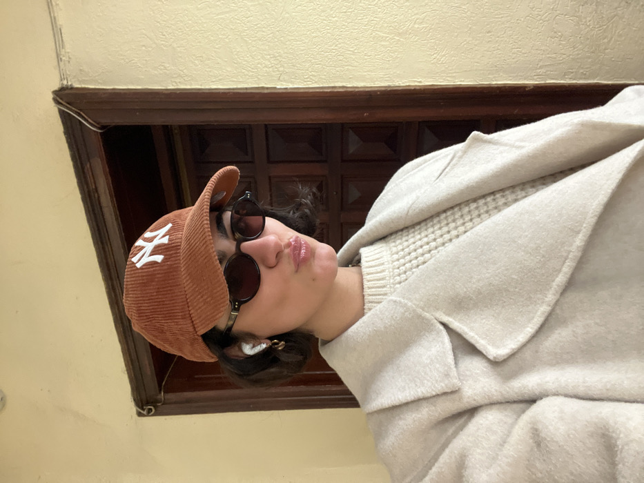 A young adult female is depicted in a close-up, head-and-torso selfie, posing with pursed lips. She wears a rust-orange corduroy baseball cap with a white "NY" logo, dark oval sunglasses, and white wireless earbuds in both ears. Small gold-colored hoop earrings are visible. Her attire includes a light beige chunky knit sweater beneath a light beige wool overcoat with visible lapels. The background consists of a plain, light beige wall and a dark brown wooden door frame leading to a paneled wooden door. A thin white cable runs along the top right corner where the wall meets the door frame. This scene is located in the city of Paris, France.