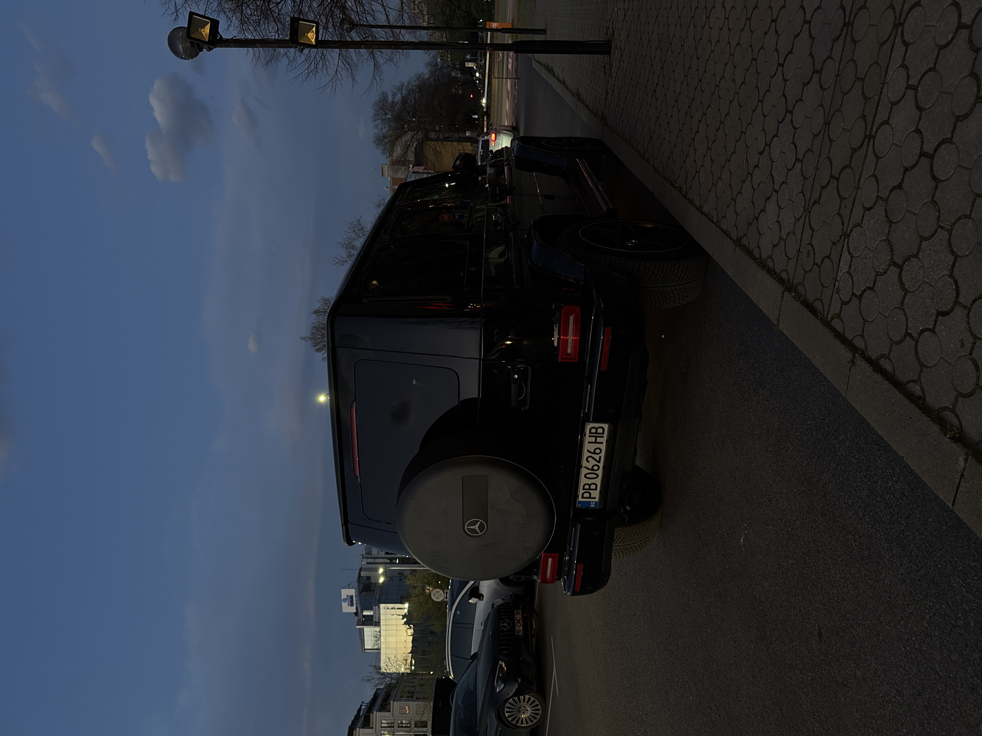 A black Mercedes-Benz G-Class is parked on the side of a street in Plovdiv, Bulgaria. The license plate reads "PB 0626 HB". Behind the G-Class is a black sedan with license plate "H6 CHO 99". The background includes buildings, streetlights, and trees. The time appears to be evening or dusk. The street has a brick-patterned sidewalk.