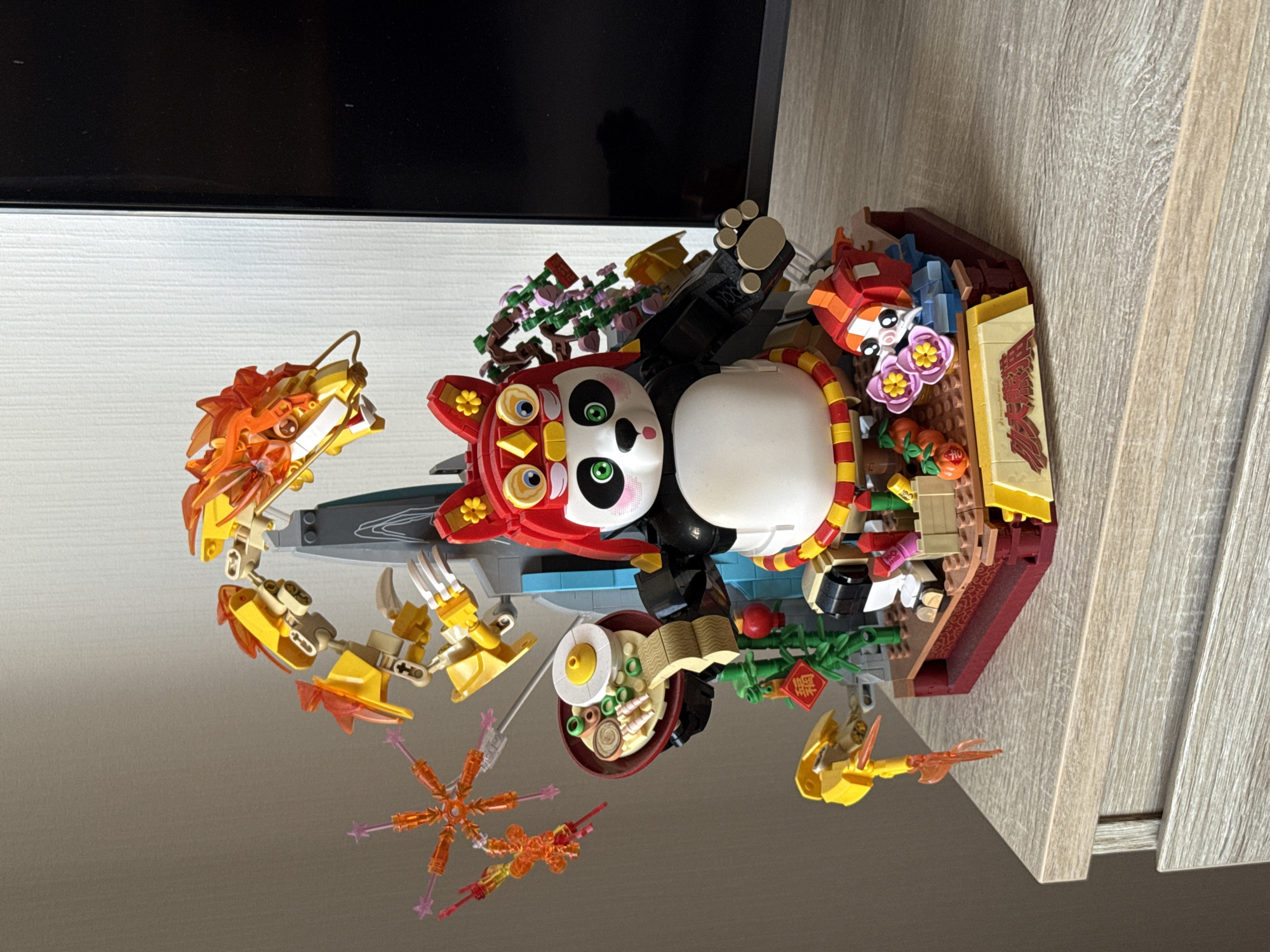 A LEGO build featuring a panda wearing a red and yellow headdress and a red and yellow sash, holding a bowl of food, stands on a red and brown base. A yellow and orange dragon figure curves above and behind the panda. To the left, an orange, star-shaped decoration hangs. Bamboo stalks with a red lantern and a red tag featuring Chinese characters are to the left of the panda. A small orange and white fish-like creature is on the right side of the base. The build is placed on a light-colored wooden surface, possibly a dresser, with a textured finish. The background consists of a light-colored wall on the left and a dark rectangular object, likely a television screen, on the right. The image appears to be taken indoors, with natural light coming from the left.