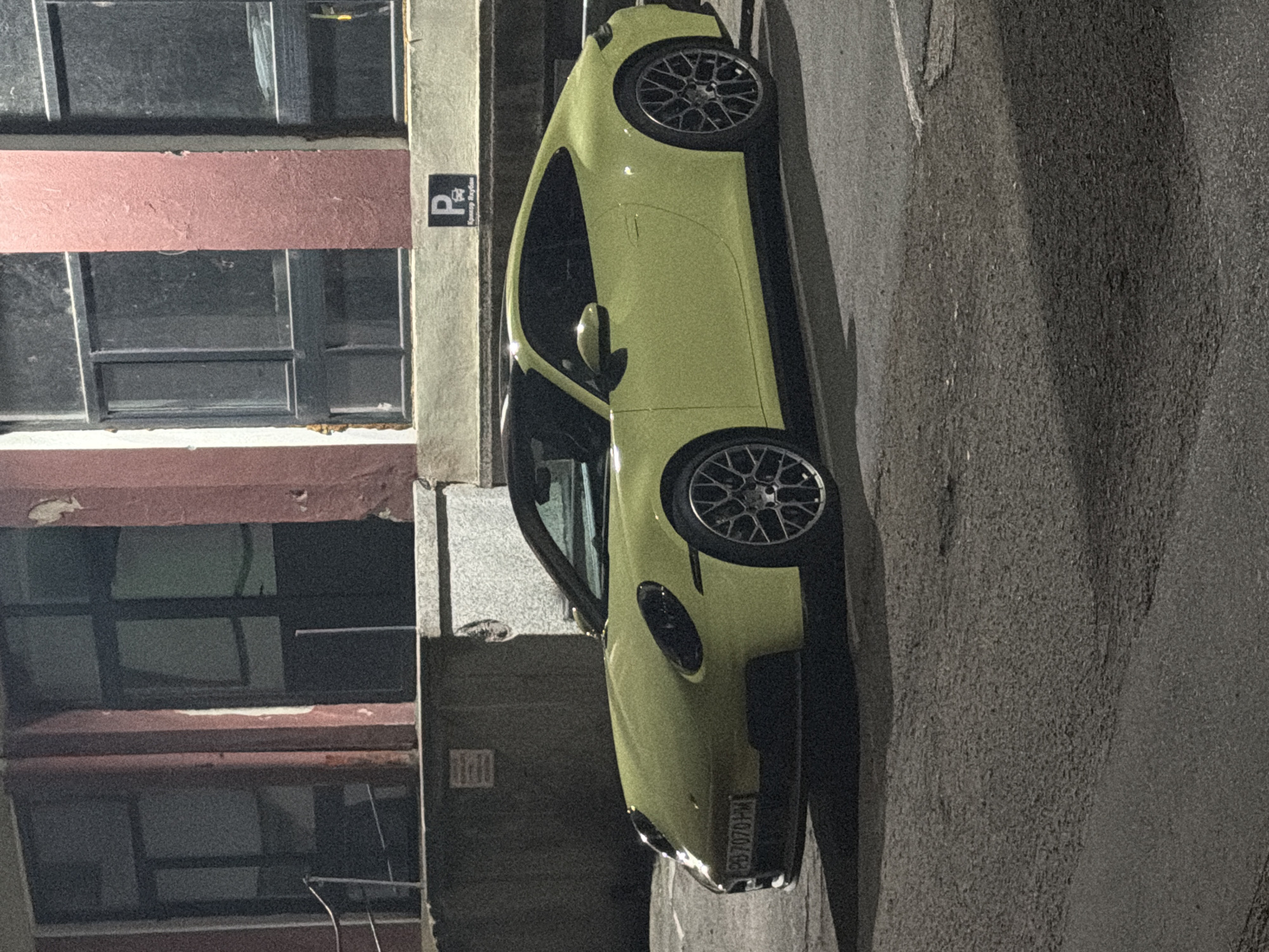 A light yellow Porsche 911 is parked on an asphalt surface in front of a building. The building has a pink facade with large, dark-paned windows and a concrete base. A white and black parking sign with the letter "P" and a car symbol is affixed to the building wall above and to the right of the car. The license plate of the Porsche reads "PD 7070 HM". The scene appears to be in an urban environment at night. The location is Plovdiv, Bulgaria.