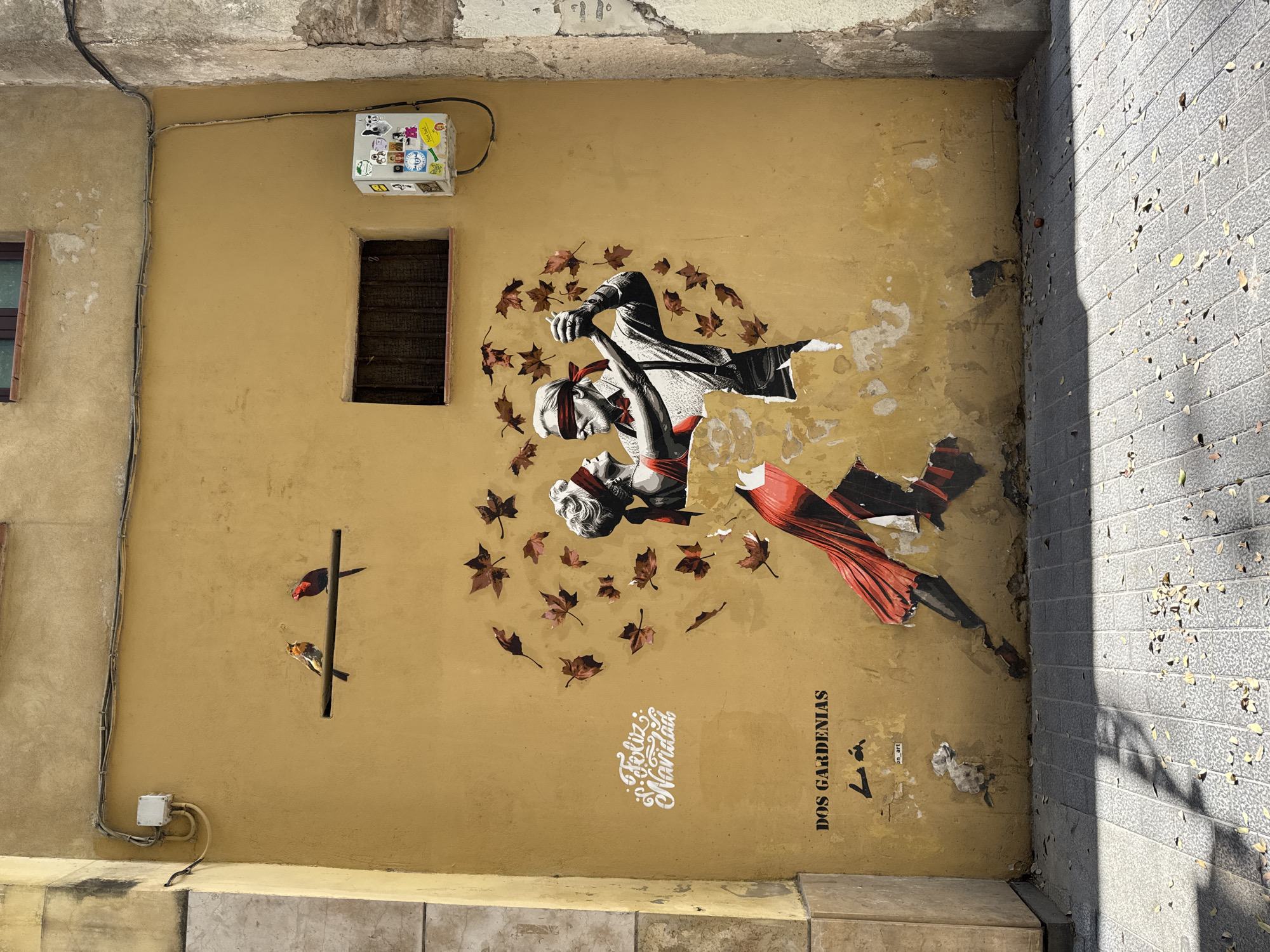 Here is a detailed description of the image:
This is a photograph of a wall in Barcelona, Spain, featuring street art. The wall is painted a yellowish-beige color and has a rough, slightly weathered texture. Affixed to the wall is a mural depicting two figures, seemingly dancers, surrounded by falling leaves. The figures have a black and white base with splashes of red, including a red blindfold on one figure. The overall impression is dramatic and artistic.
To the left of the mural, there is a dark metal pole with two small birds perched on it. Electrical cables and boxes are mounted on the wall above and below the birds. There's a small, rectangular opening with a dark grate nearby.
On the lower right side, the wall meets a brick paved walkway which is partially in shadow and covered with leaves. The lower portion of the wall near the walkway appears worn and torn, showing the effects of exposure to the elements.
Text elements in Spanish are present on the lower right of the wall next to the dancers, including "Feliz Navidad," "Dos Gardenias," and the on_art marker.