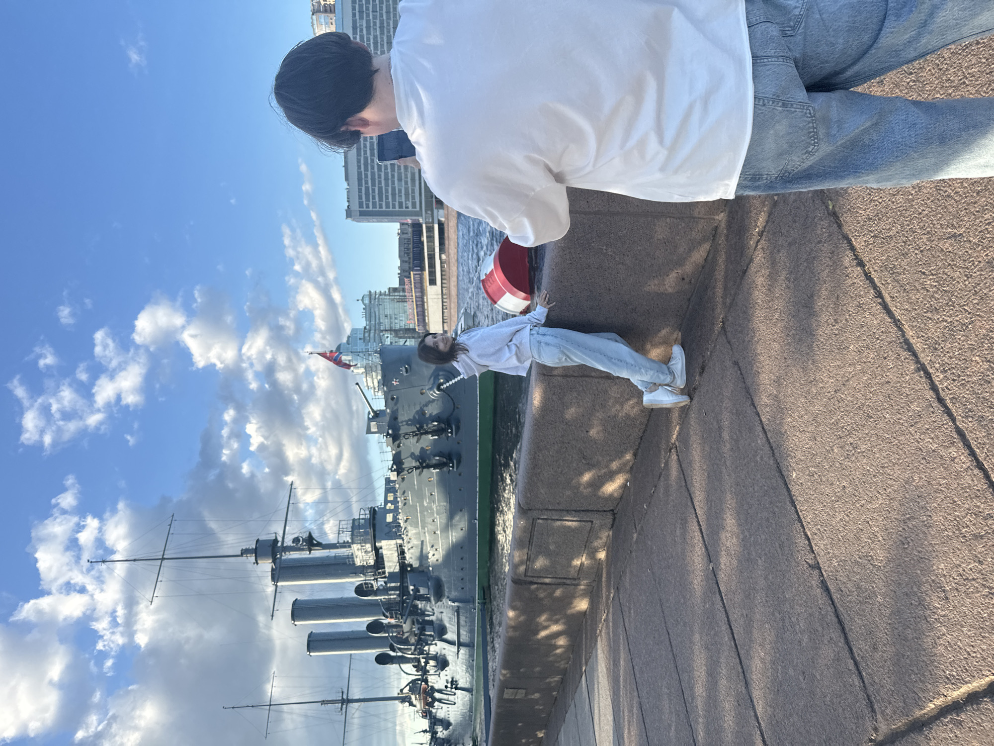 In Saint Petersburg, Russia, a young woman in a light-colored hoodie and jeans poses for a photograph taken by a young man wearing a white t-shirt and jeans. The woman stands on a stone embankment, her body angled towards the camera, looking over her shoulder. Behind her, a large, historic warship with multiple smokestacks and masts is docked. The ship is dark gray with a green waterline and appears to be a museum or monument. A red and white buoy is also visible near the woman. The scene unfolds under a bright blue sky dotted with fluffy white clouds, with buildings visible in the background beyond the ship. The ground in the foreground is paved with large, irregular stone slabs, casting long shadows. The sun is low in the sky, suggesting it is late afternoon or early evening.