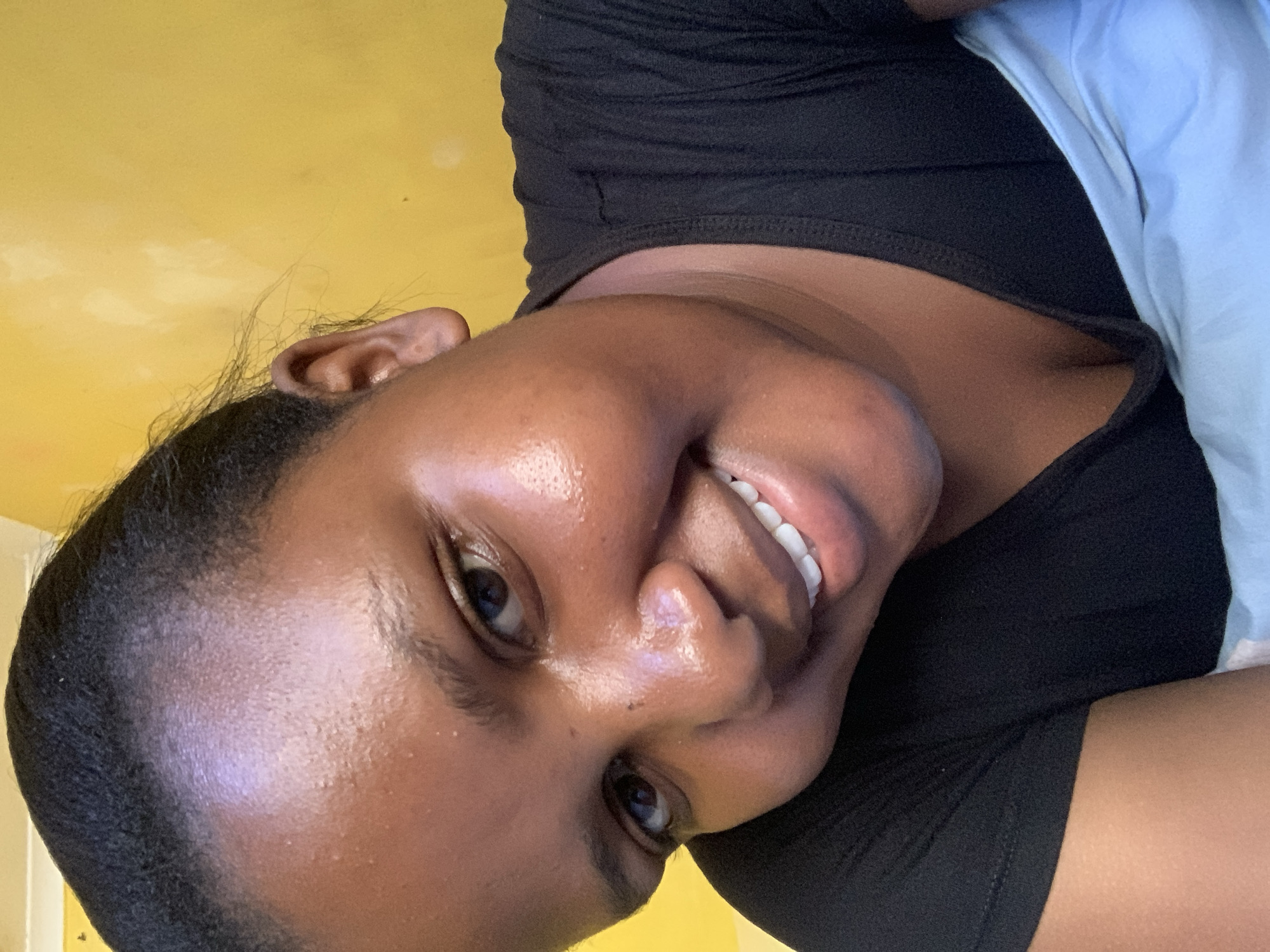 An eye-level, close-up, portrait of a young Black woman is captured indoors under natural lighting with a yellow wall in the background. The woman is smiling, exposing her upper teeth. She is wearing a black shirt with a hint of light blue fabric at the edge of her right shoulder. Details about her location in Dar es-Salaam, Tanzania are not provided in this image.