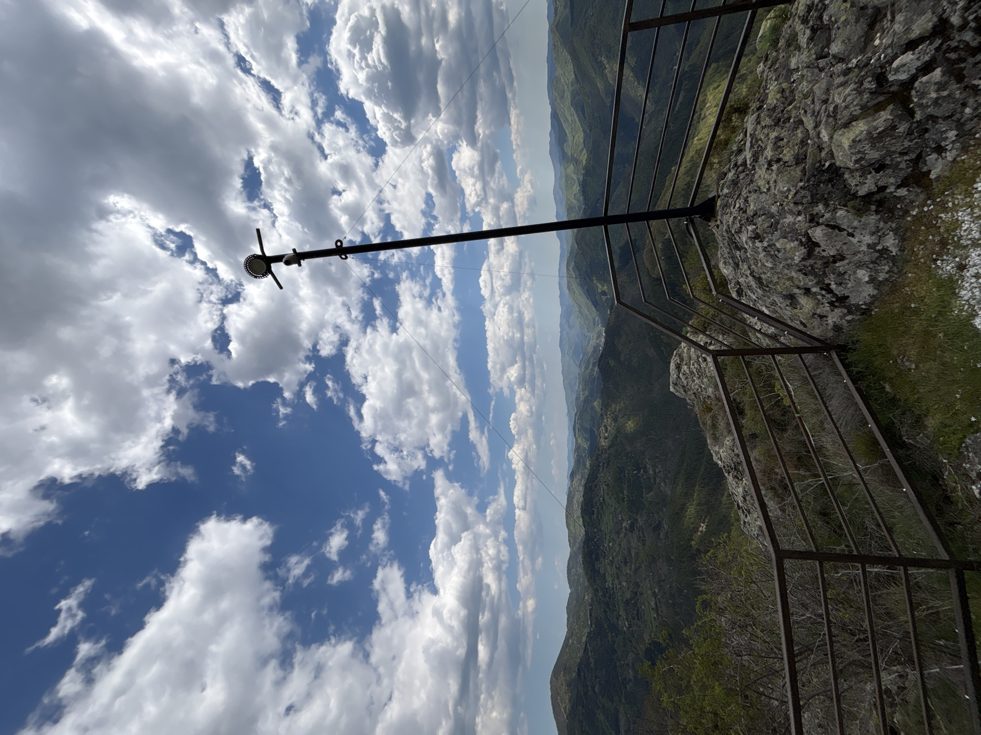 The image shows a view from a cliffside in Vlahovo, Bulgaria. A metal safety railing is present on the cliff edge. A pole extends upwards, supporting what appears to be a surveillance camera and a light fixture. A wire stretches horizontally from the pole. The background shows a landscape of rolling hills and valleys. The sky above is filled with white clouds.