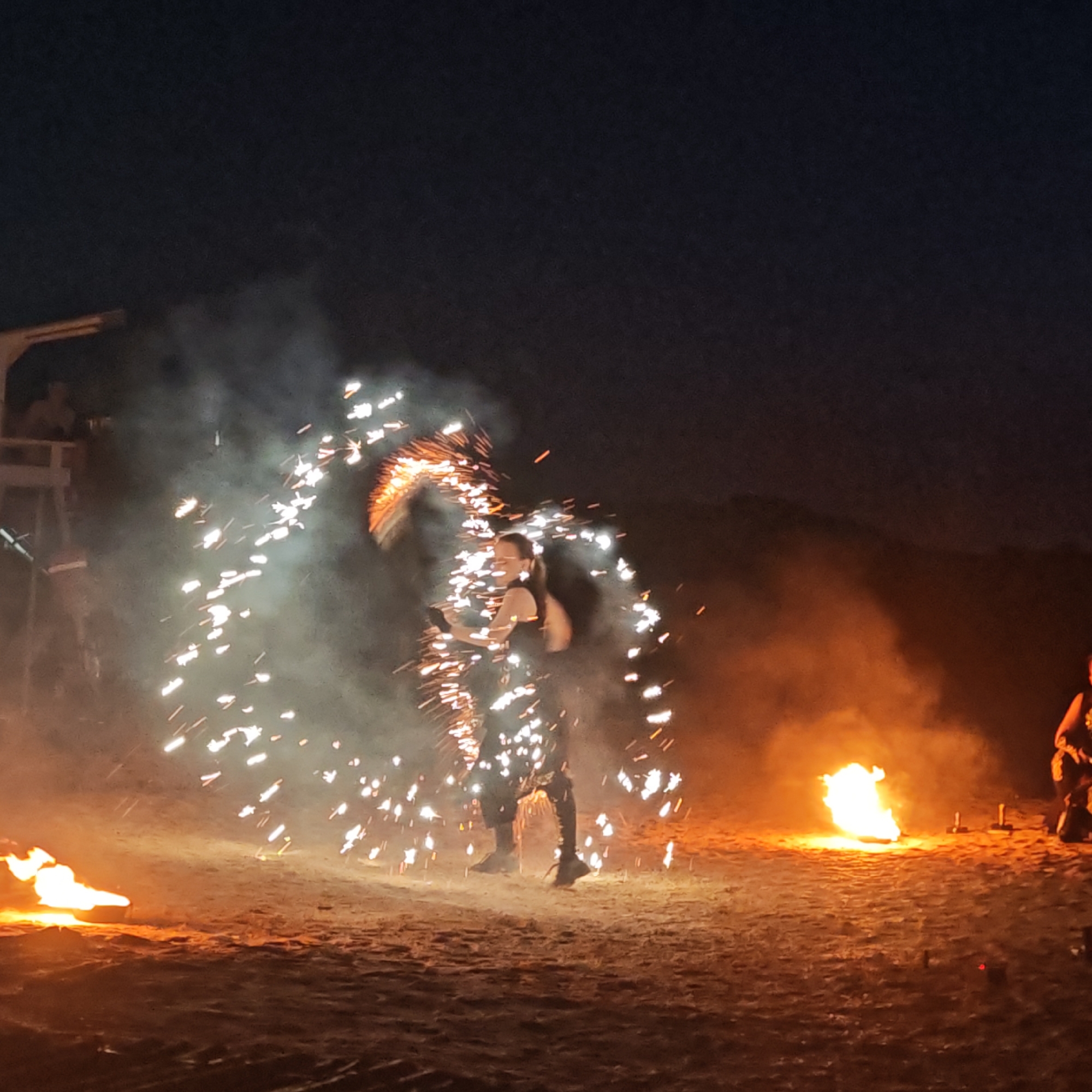 An outdoor nighttime performance is underway in Mysivske rural hromada, Ukraine, featuring a fire dancer. The performer, dressed in dark clothing, is actively twirling a flaming prop that emits a shower of sparks, creating a dramatic visual effect against the dark sky. Smoke billows around the performer, illuminated by the bright sparks and the intense glow of several large bonfires positioned on the sandy ground. To the left, a wooden structure, possibly a stage or viewing platform, is visible with a silhouetted figure standing on it. The overall scene is one of dynamic action and fiery spectacle.