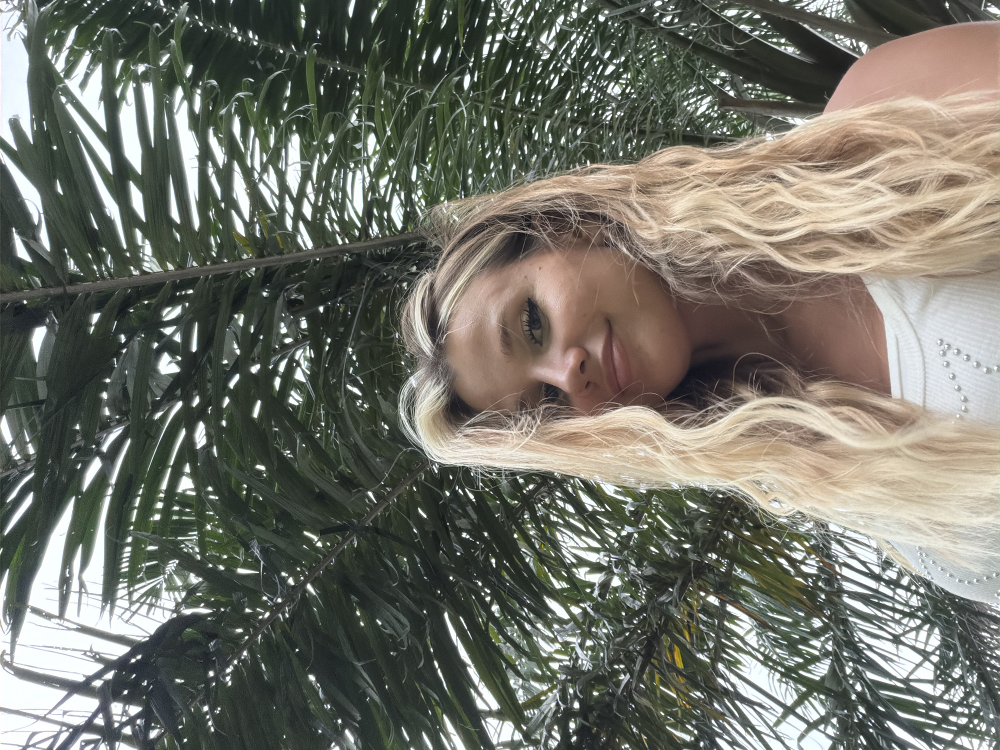 A young woman with blonde, wavy hair is captured in a low-angle, outdoor selfie. She is positioned centrally in the frame, looking slightly off to the side with a gentle smile. She is wearing a white, sleeveless top adorned with small, shimmering embellishments. Behind her, a dense canopy of palm fronds creates a lush, green backdrop against a bright, overcast sky. The scene suggests a tropical or subtropical environment, possibly within the vicinity of Choeng Thale, Thailand, known for its beaches and palm-lined landscapes.