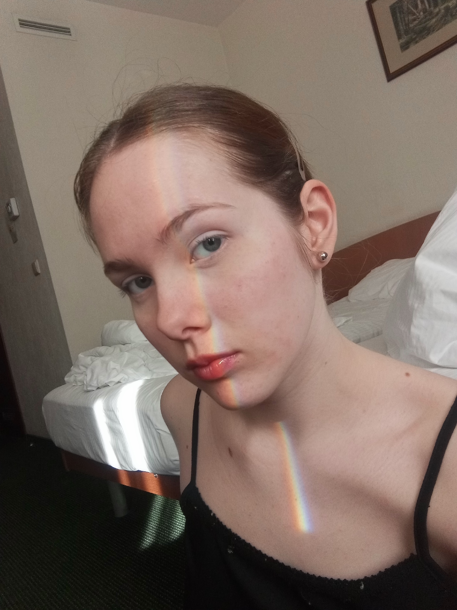A fair-skinned young woman with light reddish-brown hair pulled back poses for a selfie. A beam of sunlight refracted into a rainbow falls across her face and chest. She has light eyes and a serious expression. She is wearing a black camisole. Behind her is a made bed with white sheets and pillows. In the background is a wall with a framed picture and an air conditioning vent. The presence of a hotel bed, drab green carpet, and generic art suggest this photo was taken in a modest hotel room. The architecture and interior are consistent with many hotels found in Saint Petersburg, Russia.