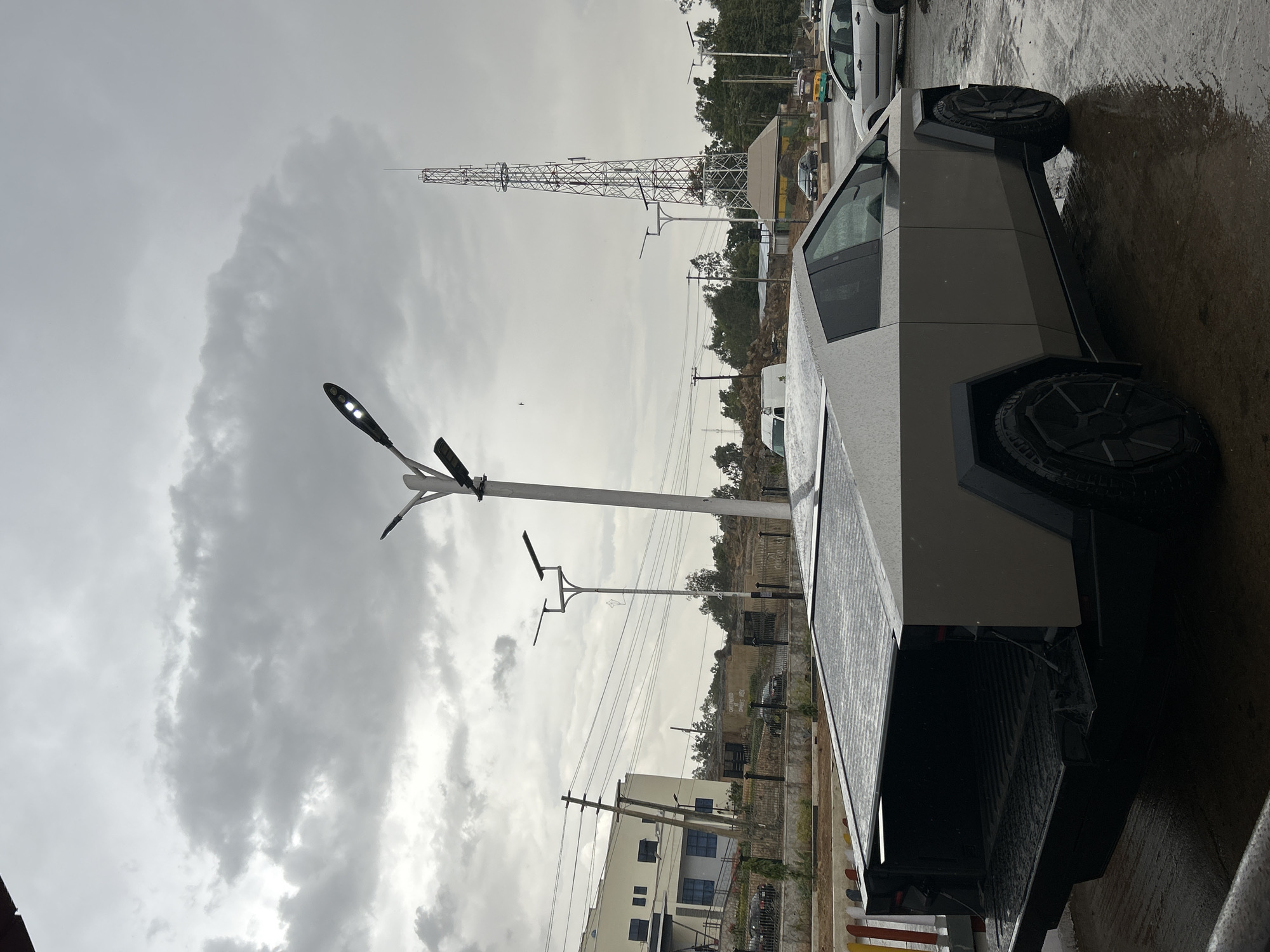 The image depicts a gray Tesla Cybertruck parked on a wet surface, likely pavement, in Jos, Nigeria. In the background are utility poles and buildings, including one with multiple windows. A tall metal lattice tower is visible, characteristic of telecommunications infrastructure. Above, overcast skies suggest recent or ongoing rainfall. A modern streetlight stands near the Cybertruck.