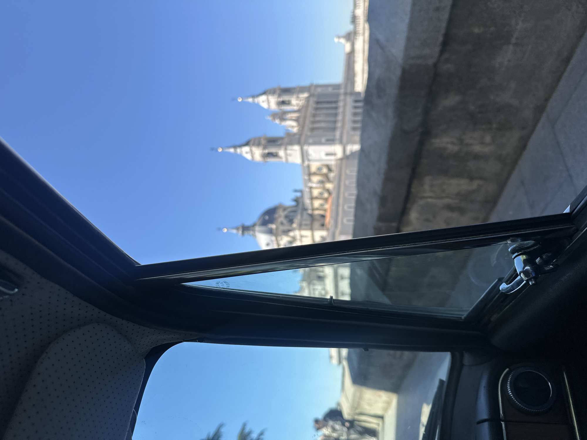 An upward-angled shot captures a majestic white architectural landmark against a clear, bright blue sky, viewed from inside a car. The building, likely a cathedral or palace, features multiple spires and domes, with its intricate details slightly blurred. A large, dark concrete structure, possibly a bridge or overpass, runs along the right side of the frame, partially obscuring the landmark.
The interior of the car is visible in the foreground, including a grey, perforated headliner on the left and the dark frame of an open sunroof. A metallic latch mechanism for the sunroof is visible on the right, above a portion of the car's door panel or dashboard with a circular vent or control. Sunlight illuminates the scene, indicating a clear, sunny day. Faint reflections, possibly of a person or trees, can be seen in the car's window. The architectural style suggests a European setting, and with the provided context, the landmark is in Madrid, Spain. Minor, illegible text appears on the car window glass.