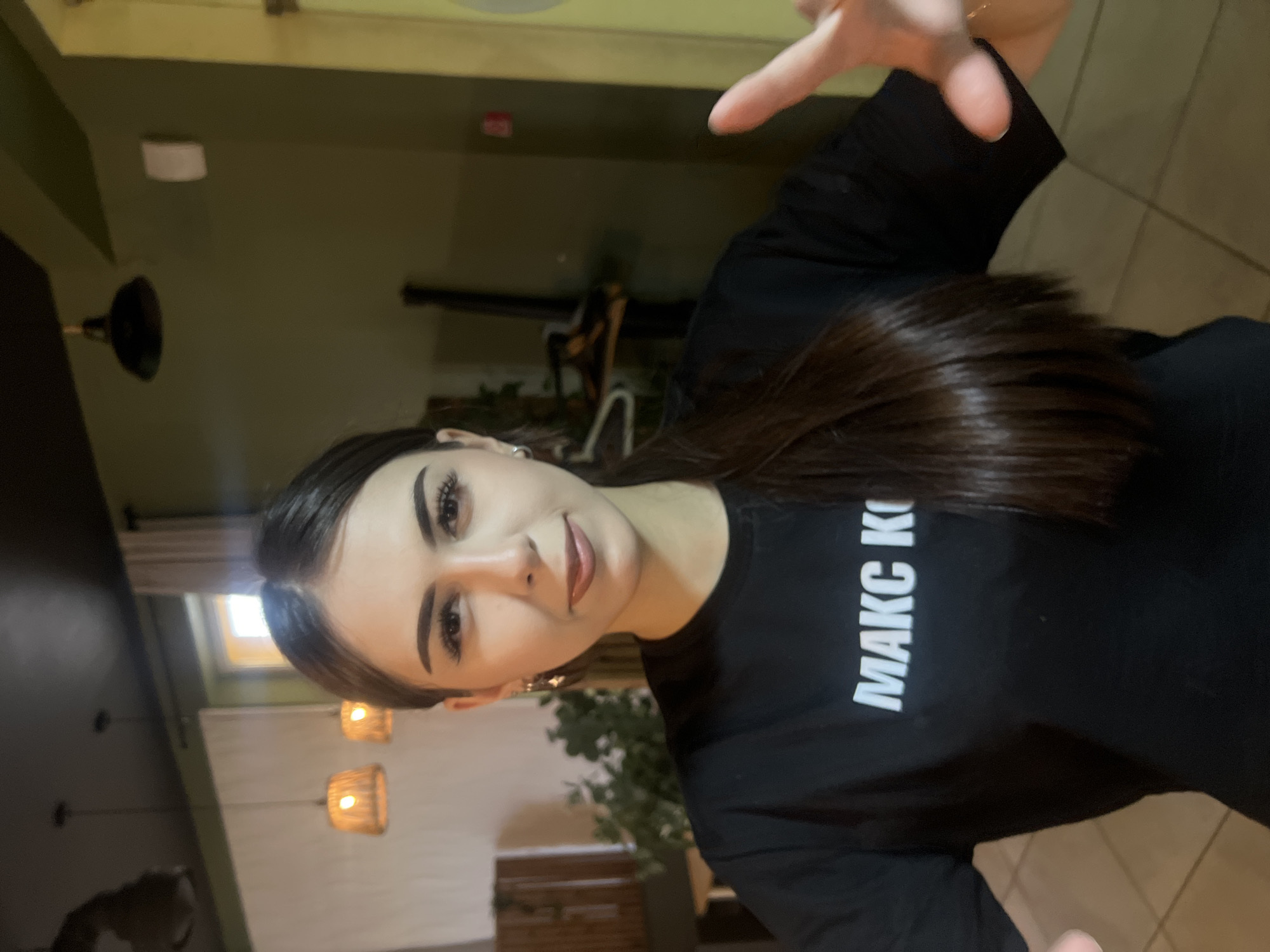 A young adult female is centrally positioned in a medium indoor shot, captured in what appears to be a cafe or restaurant located in Красные Баки, Russia. She has long, straight, dark brown hair parted in the middle and styled back, revealing small silver-colored hoop earrings. Her face features defined eyebrows, eyeliner, mascara, and brownish-mauve lipstick. She wears a black short-sleeved t-shirt with the white Cyrillic text "МАКС К" printed across the chest. She looks directly at the camera with a neutral expression, while her right hand is partially extended towards the camera with fingers slightly spread.
The background is dimly lit and slightly out of focus. It features a dark ceiling from which two woven pendant lights and one dark industrial-style pendant light hang. The left wall appears light-colored, possibly with a textured panel, and includes dark wooden paneling and some green foliage. The right wall is olive green and has a small, rectangular light-colored fixture mounted on it. The floor visible at the bottom of the frame is tiled in a light brown hue.