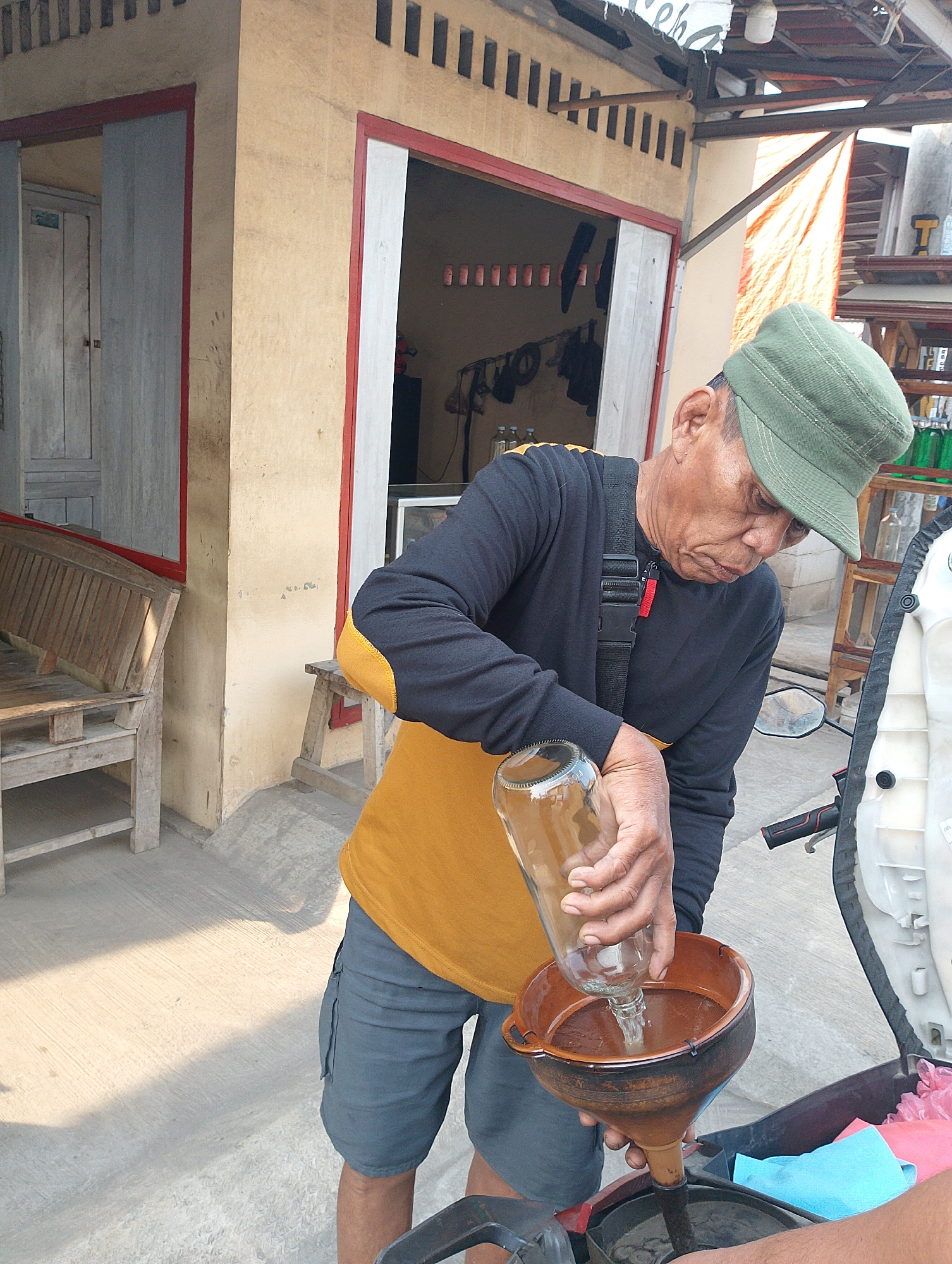 In Karawang, Indonesia, an elderly man wearing a green cap and a black long-sleeved shirt over an orange t-shirt and grey shorts is pouring liquid from a glass bottle into a brown funnel, which is positioned over a motorcycle. The scene takes place outdoors, with a rustic building featuring yellow walls and red trim in the background. A wooden bench is visible to the left, and a wooden stall with shelves is to the right, displaying several green bottles, possibly containing liquids. The ground is paved with concrete, and sunlight casts shadows. The man appears focused on the task of pouring the liquid, suggesting he might be refueling or adding a substance to the motorcycle.