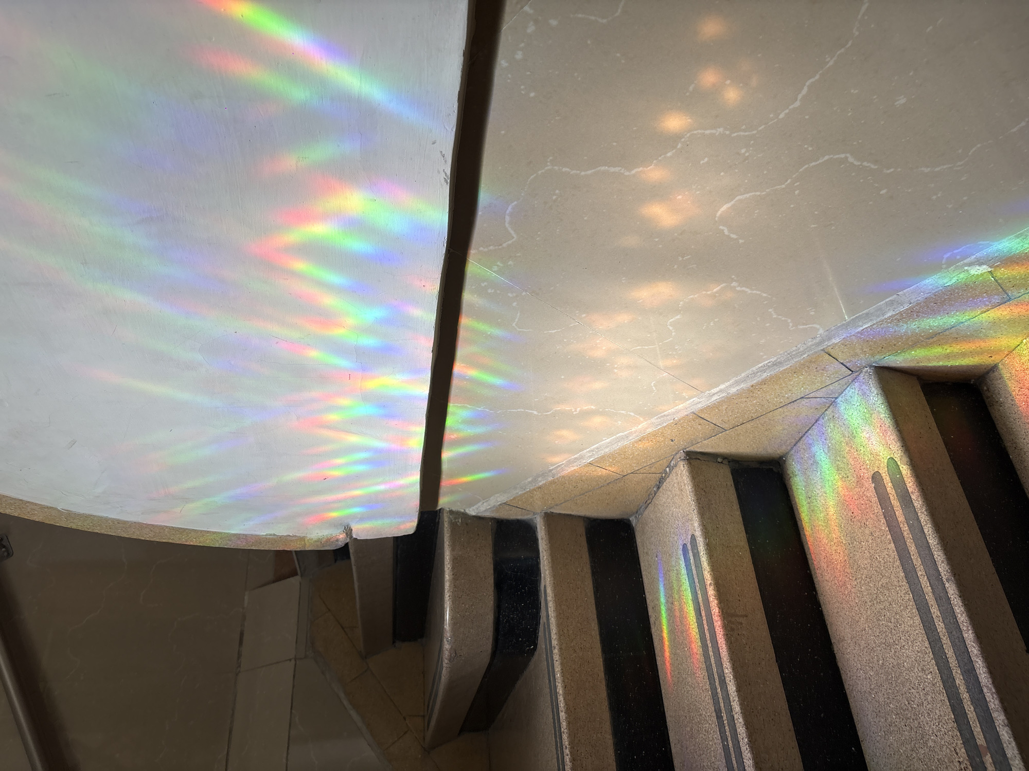 An overhead shot captures a winding staircase illuminated by vibrant, multi-colored light reflections in an interior setting in Mexico City, Mexico. The staircase features curved steps with textured, beige-colored risers and treads, each marked with two parallel, dark grey or black anti-slip strips. These strips create a striking contrast against the lighter stone.
The walls and underside of the upper landing, which are light beige or off-white, are bathed in brilliant prismatic light, forming elongated, wavy rainbow patterns. These colorful reflections, displaying hues of red, orange, yellow, green, blue, and violet, also extend onto the steps of the staircase. The light source, likely natural sunlight passing through a window or object that refracts light, suggests it is daytime. The surfaces of the walls and ceiling show some subtle texture and faint crack patterns. No people are visible in the scene, and there is no discernible text. The overall atmosphere is bright and visually dynamic due to the colorful interplay of light and architecture.