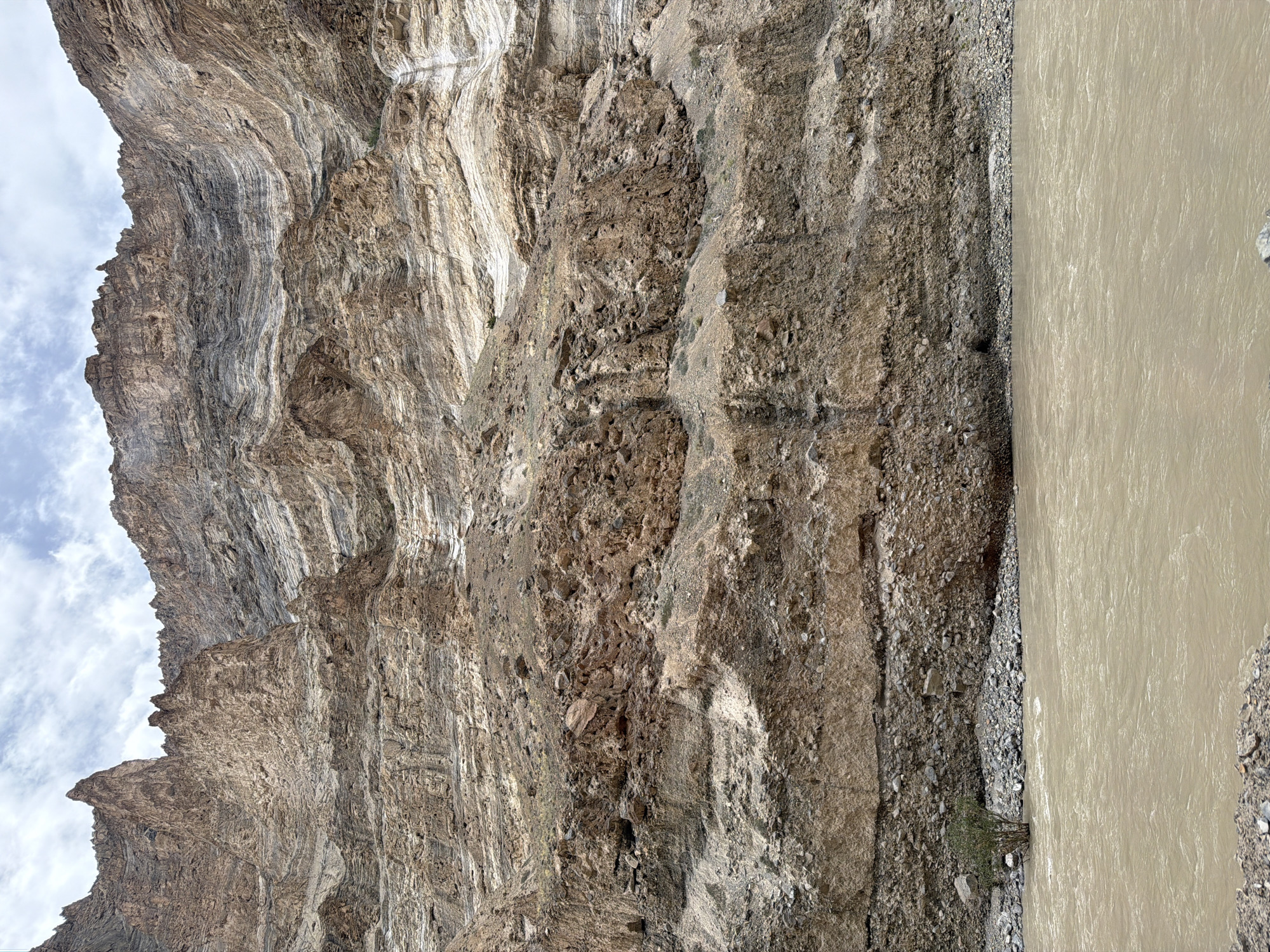 The image shows a wide, muddy river flowing in front of a massive, layered cliff face. The cliff is composed of various shades of brown, grey, and white rock, with distinct horizontal striations indicating geological stratification. The terrain at the base of the cliff is rocky and barren, with a single small, scrubby tree visible on the left side of the riverbank. The sky above is overcast with patches of white clouds. This scene is characteristic of the landscape found near Stongde, India, a region known for its dramatic mountainous terrain and glacial rivers.