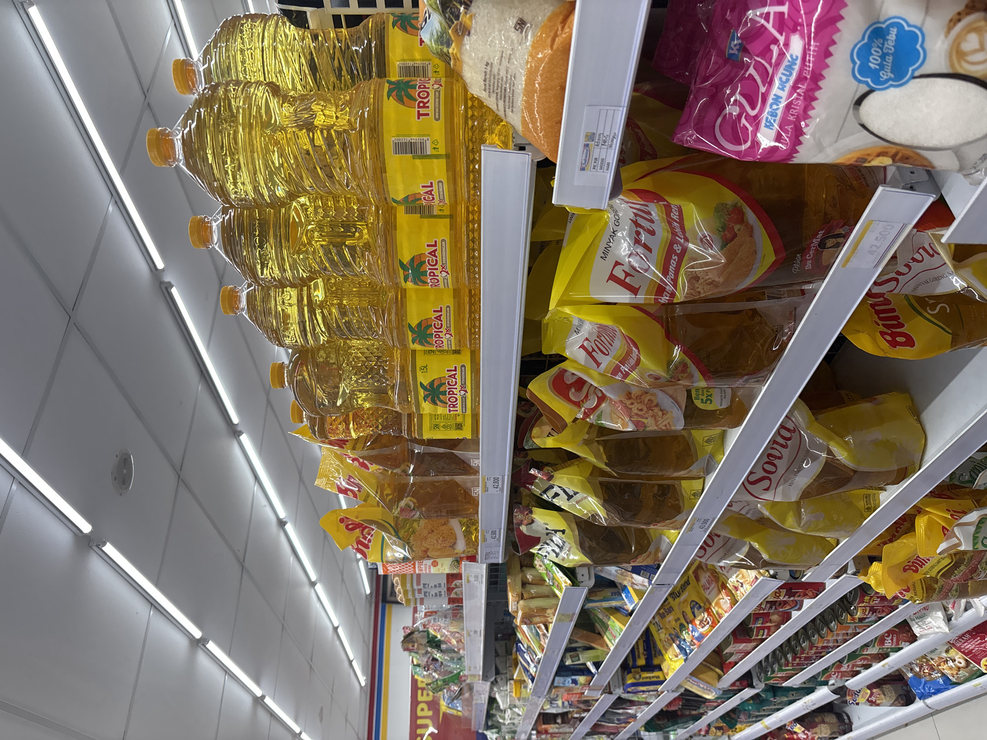 The image shows an aisle in a supermarket in Cirebon, Indonesia, stocked with various food items. The focus is on the shelves, which are filled with cooking oil in large plastic bottles and pouches, as well as bags of sugar and other packaged goods. The lighting appears to be from fluorescent tubes in the ceiling. The shelves are organized with products from brands like "Tropical," "Fortune," and "Sotra." There are price tags visible on some of the shelves. The overall impression is of a well-stocked retail environment.