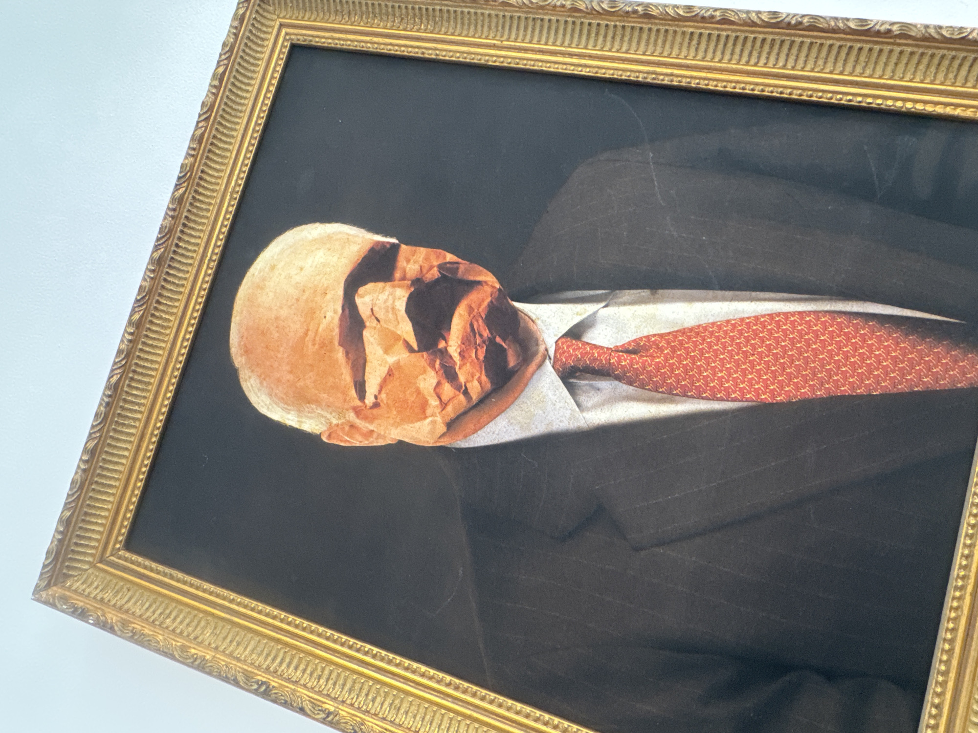 A framed artwork of a man in a suit with a red tie hangs on a white wall. The man's head appears to be made of crumpled paper. The frame is gold and ornate. The location, Tel-Aviv, Israel, is not discernible from the image.
