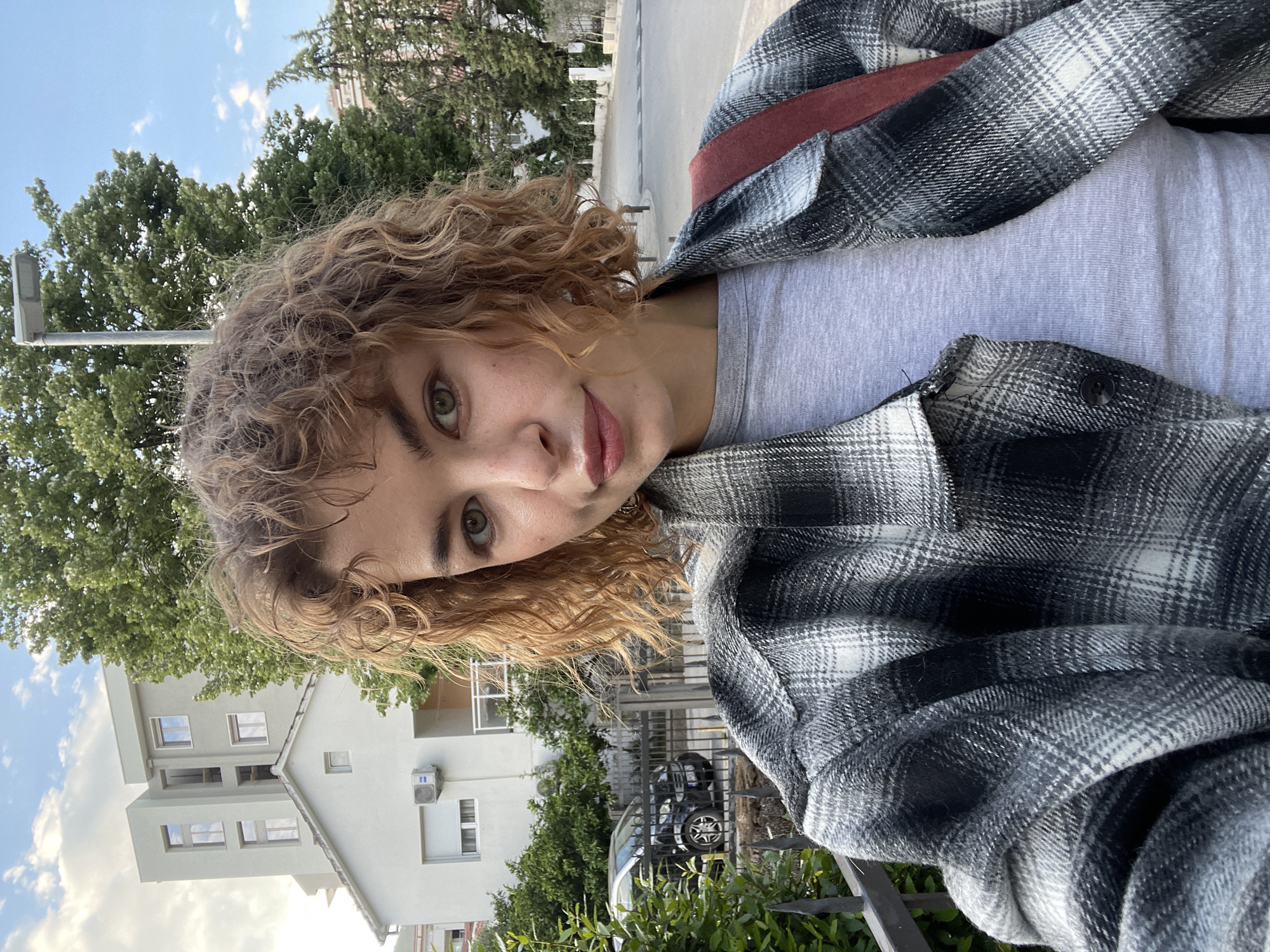 A young person with curly brown hair and green eyes is posing for a selfie. They are wearing a grey t-shirt and a black and white plaid flannel shirt. A red backpack strap is visible over their shoulder. The background includes a building with an air conditioning unit, trees, a lamppost, and a car parked behind a fence. The sky is blue with some clouds. The location appears to be Budva, Montenegro, based on the architecture and vegetation visible in the background.