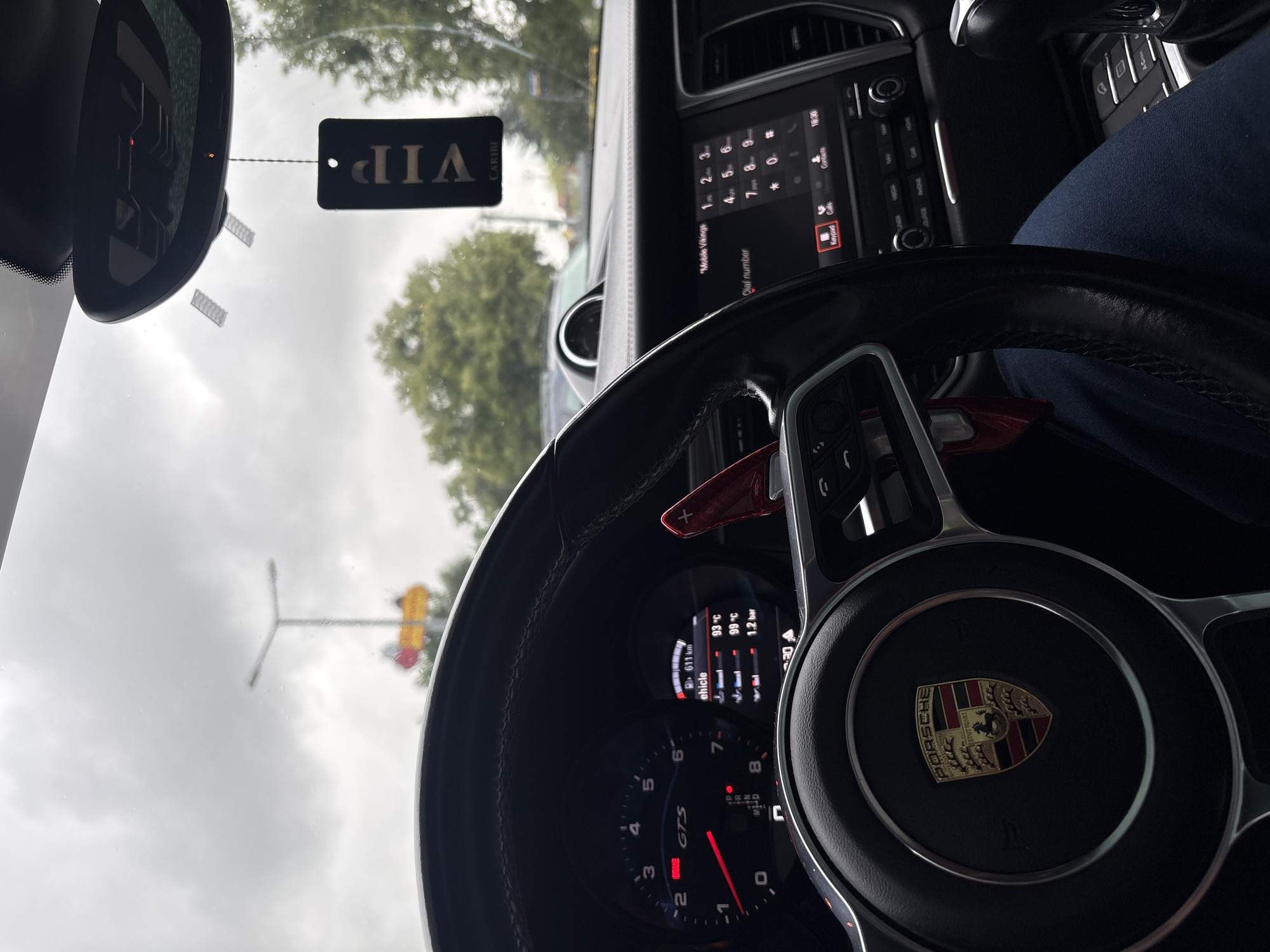 The image provides an inside view of a car, likely a Porsche, on a cloudy day in Wydminy, Poland. The focus is on the steering wheel and dashboard. A "VIP" tag hangs from the rearview mirror, suggesting a special occasion or status. The dashboard displays various information including vehicle speed (indicated by "GTS"), engine temperature (93°C and 99°C), and tire pressure (1.2 bar). The infotainment system is visible, showing a phone dial pad with "Mobile Vikings" and "CARIBI" text. The time displayed is 16:30, indicating late afternoon. Outside the car, a traffic light and trees are partially visible through the windshield, suggesting the car is stationary, possibly at a traffic light or in traffic. The overcast sky implies a likely chance of rain. The image is taken from the driver's perspective.