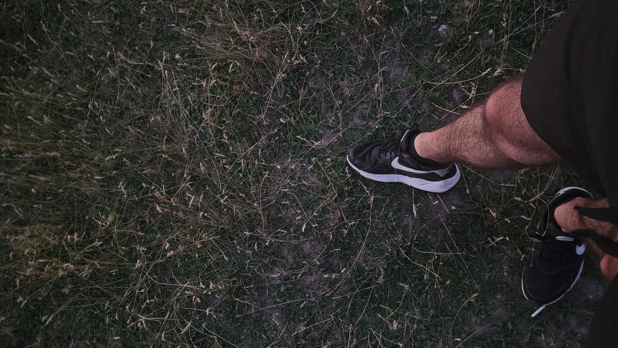 A close-up, overhead shot shows a person's legs and feet against a background of dry grass and patchy soil. The person is wearing black athletic shoes with white accents and black shorts. One foot is positioned slightly forward, while the other is turned outwards, its shoe partially obscured by the person's leg. The leg is covered in light brown hair, and its musculature is visible. The overall scene appears to be outdoors, possibly in a field or park, with the lighting suggesting either early morning or late evening.