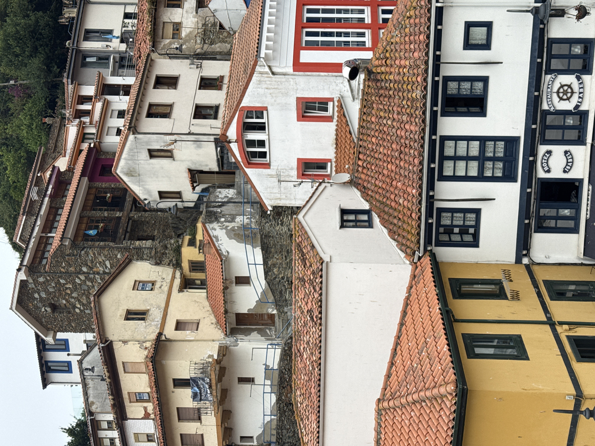 The image shows a view of the colorful houses and rooftops of Cudillero, Spain, a picturesque fishing village on the Asturian coast. The houses are built on a steep hillside, creating a tiered effect with narrow streets and alleys winding between them. The architecture is traditional, with white-washed walls, red tile roofs, and wooden shutters.
In the foreground, a yellow house with dark green trim and windows is visible. Next to it is a white building with dark blue trim and windows, featuring signs that read "HOTEL ISABEL" and "RESTAURANTE ISABEL" with a ship's wheel emblem. Above these buildings, more houses cascade up the hill, some with stone facades and others painted in various shades of white and cream, accented with red and pink. The overall impression is one of a charming, densely packed coastal town.
