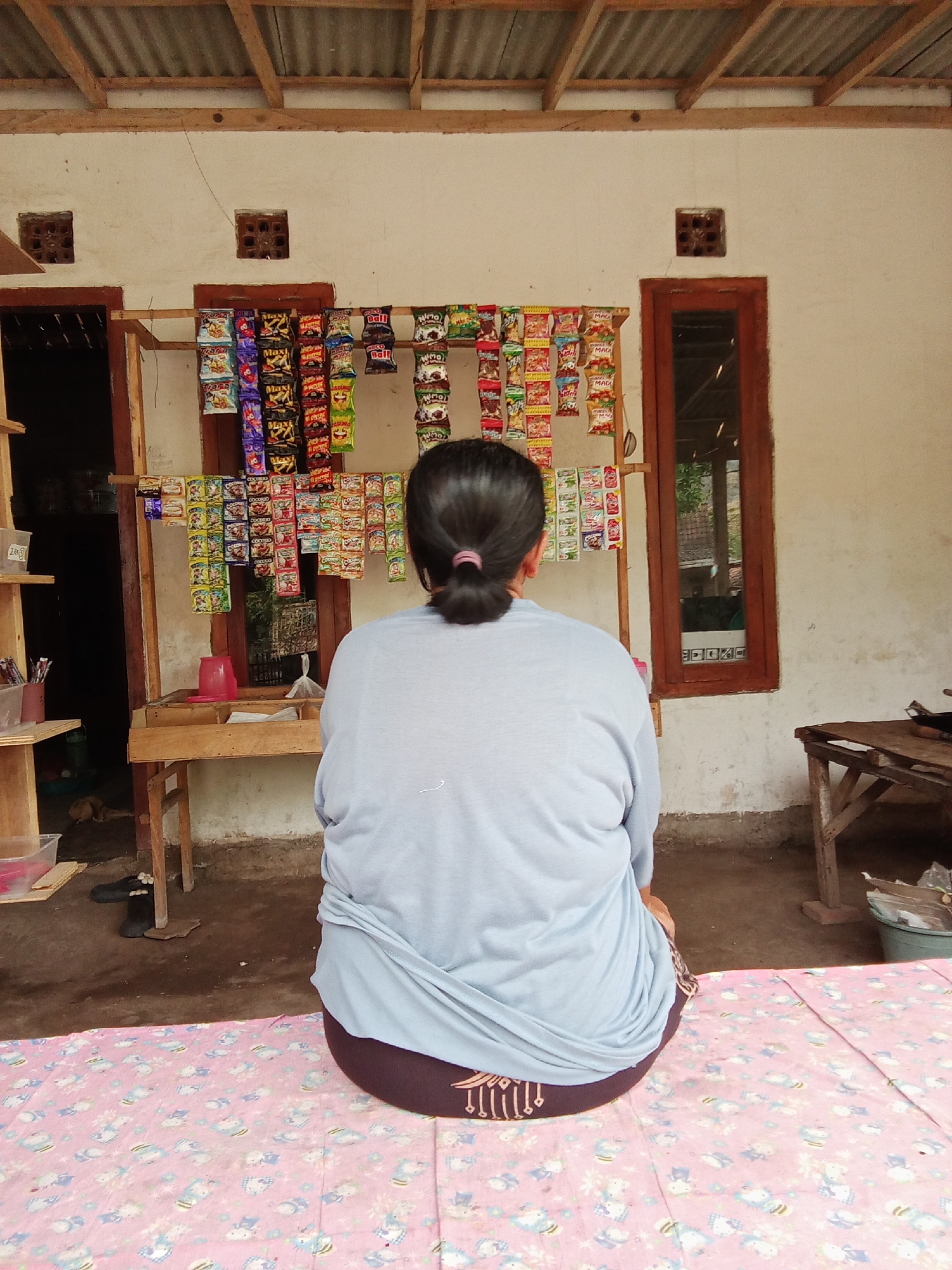 A woman sits facing away from the camera, looking at a small shop stall. The stall displays numerous small packets of snacks and candies, organized on wooden racks. Behind the stall, the wall of a building is visible, with two small, square openings in the wall. A wooden door with a window is to the right of the snack display, and another open doorway is to the left. The ground in front of the stall is dirt, and the woman is sitting on a pink mat with a pattern of cartoon characters. The scene appears to be outdoors, in a rural or semi-rural setting, likely in Trebungan, Indonesia.