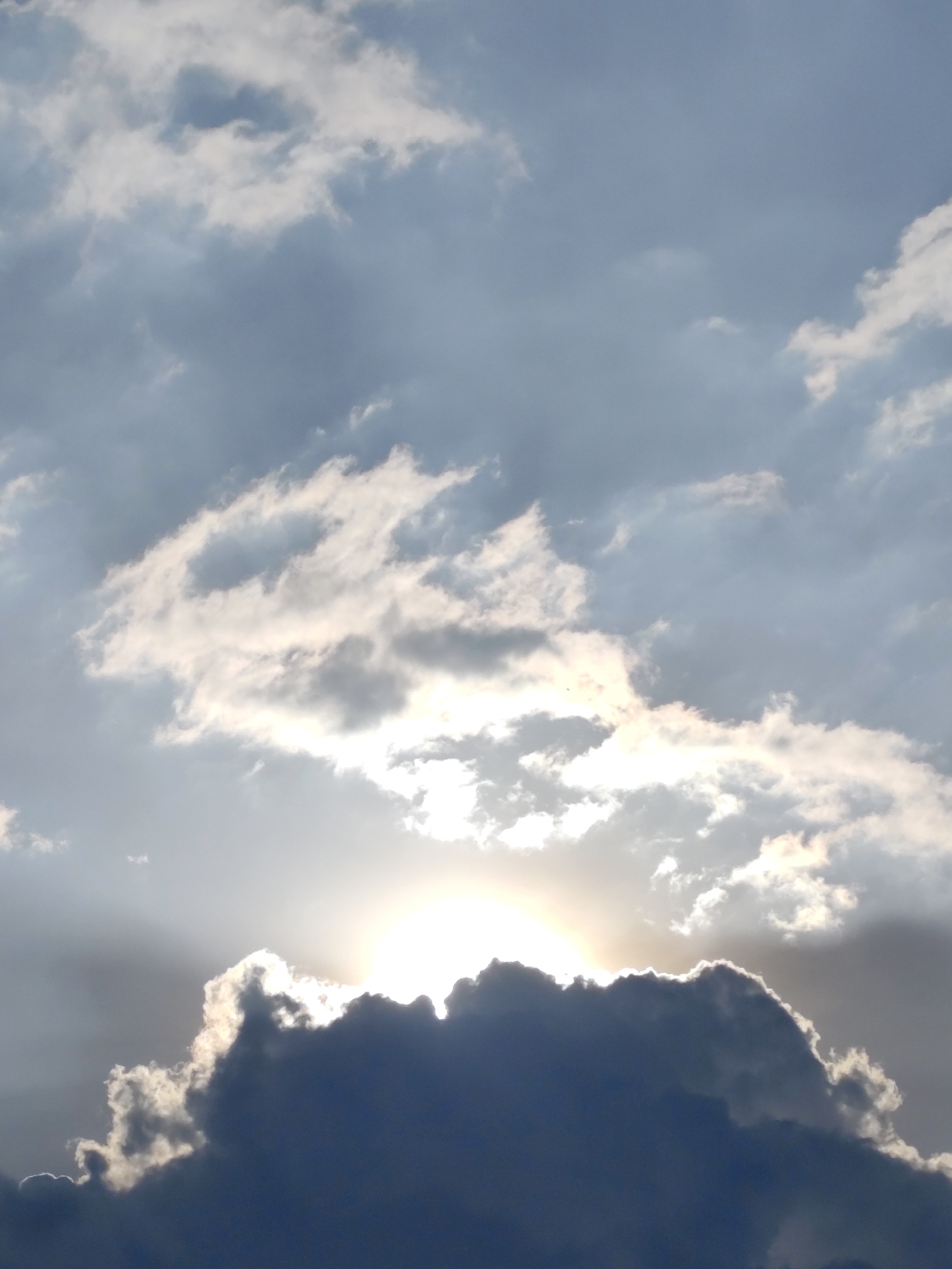 The image shows a sky filled with clouds, with the sun peeking through the clouds. The lower part of the frame is dominated by a dark, heavy cloud. The location and the city are not identifiable from the image.