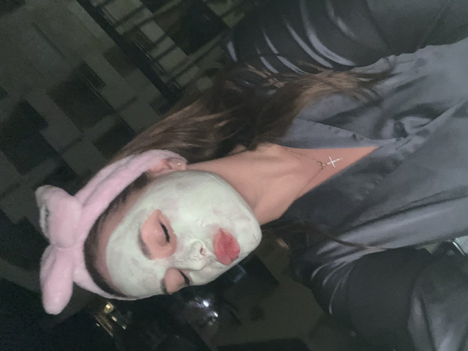 A female subject is depicted in a self-portrait taken at night in Unknown, Unknown. Her face is covered with a light green facial mask, eyes are closed, and lips are pursed. She wears a pink, plush headband with prominent bunny-ear details, and a dark gray satin robe or pajama top. A silver-toned necklace with a small, diamond-encrusted cross pendant is visible around her neck. Her long, dark brown hair is visible flowing down. The background consists of dimly lit building structures and a dark sky, consistent with an elevated outdoor setting such as a balcony.