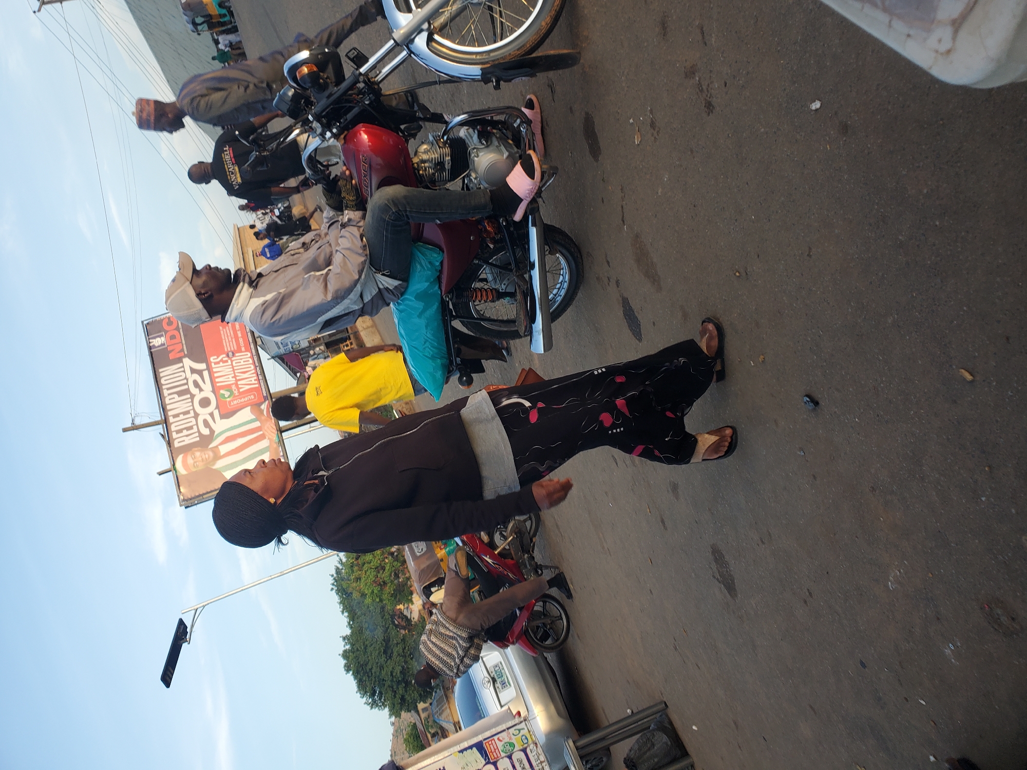 In Gwong, Nigeria, a street scene unfolds under a clear sky, where a woman in a black headscarf and jacket walks past a red motorcycle. The motorcycle is occupied by a man wearing a grey jacket and a light-colored cap, and he is looking to his right. Behind him, another man in a dark shirt and a traditional cap walks away from the camera. In the background, a large billboard displays "REDEMPTION 2027 NDC" along with a portrait of a man in traditional attire. A silver car is parked to the left of the frame. The ground is paved, with scattered debris and dark patches suggesting it is a road or a marketplace. The overall atmosphere appears to be that of a busy urban environment.