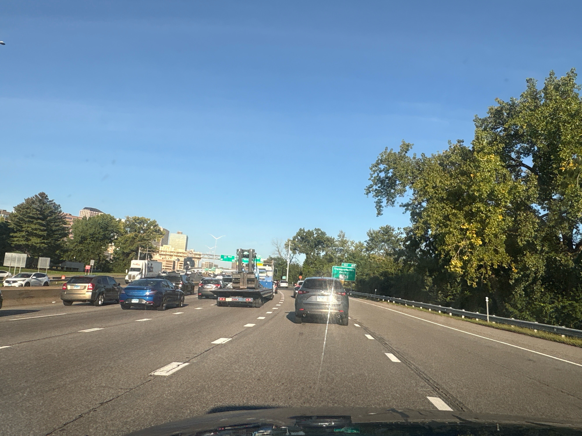 A highway scene in East Hartford, Connecticut, USA, is depicted under a clear blue sky. Multiple lanes of traffic are visible, with cars and trucks moving along the asphalt road. On the left side of the highway, trees and buildings can be seen in the distance. A white truck with a trailer is in the middle lane, while a dark-colored SUV is in the right lane. Further down the road, a dark sedan is in the right lane, and a blue sedan is in the left lane. On the right side of the highway, a dense line of trees borders the road. There are no people visible in the frame. The overall impression is a typical commute or travel on a multi-lane highway.