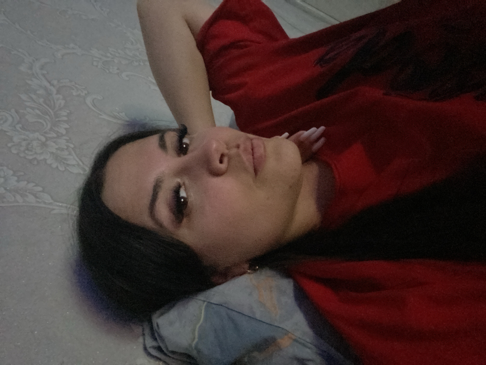 A young woman is lying down on a patterned surface, possibly a bed, with a pillow under her head. She is wearing a red top and has dark hair and visible eyelashes. Her hand is near her face. The environment appears to be indoors, but specific location information, such as Chaikovsky, Russia, cannot be determined from the image.