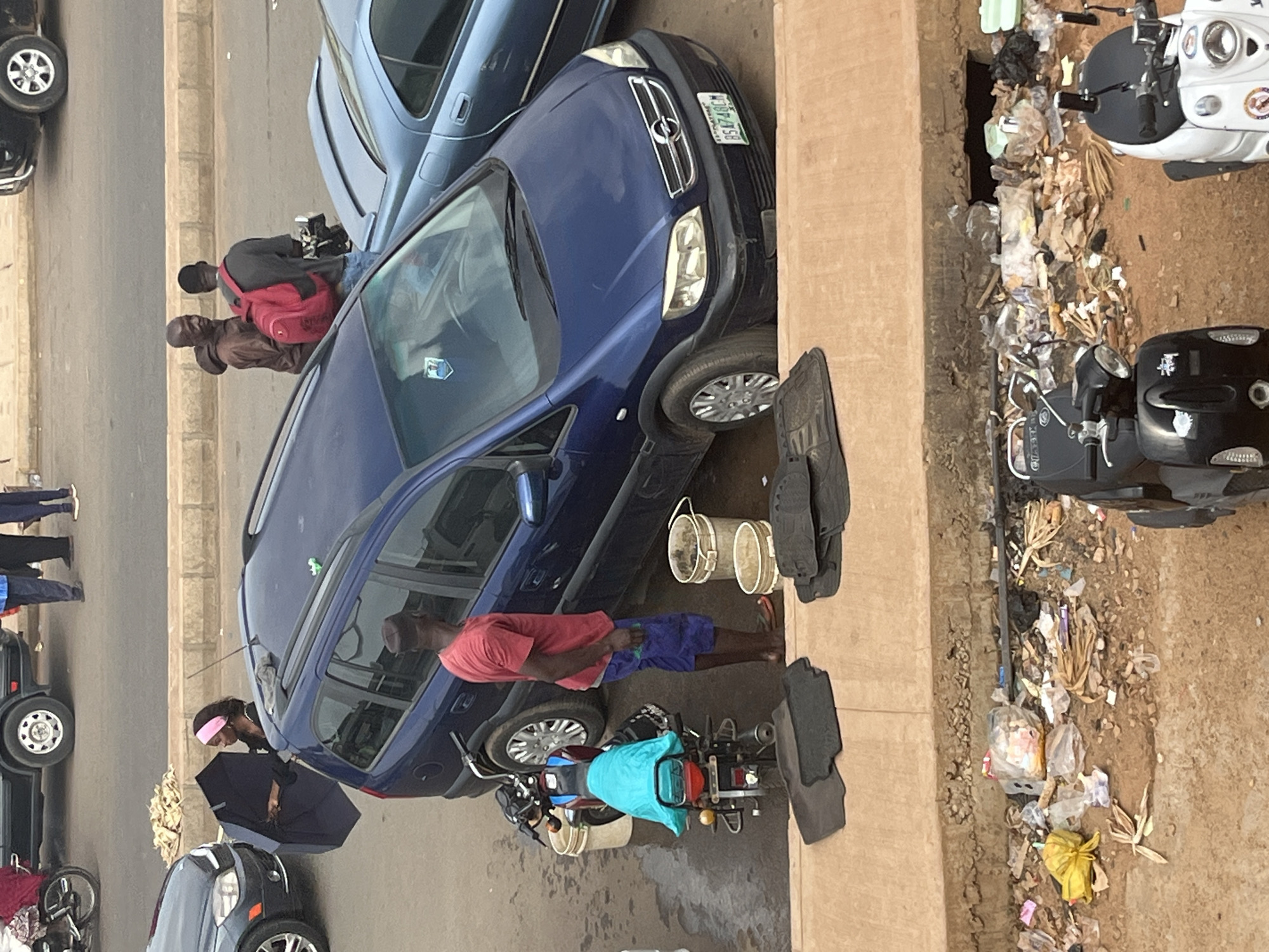 This image depicts an outdoor scene in Jos, Nigeria, likely on a street or roadside. In the foreground, there are piles of trash and debris. Two scooters are parked; one is black and the other is white. Behind the scooters, there are car mats laid out on the ground. A man in a red t-shirt and blue shorts stands next to a blue Opel Zafira minivan, which is parked on the street. Next to the minivan are several buckets, suggesting a car wash or water collection activity. A motorcycle is parked near the minivan, covered with a turquoise tarp. In the background, a woman with a pink headband is holding a dark umbrella. Two men, one wearing a red backpack, are standing near the minivan. Other vehicles, including a dark-colored car and a light blue car, are visible on the road. The environment appears to be a dusty, urban setting.