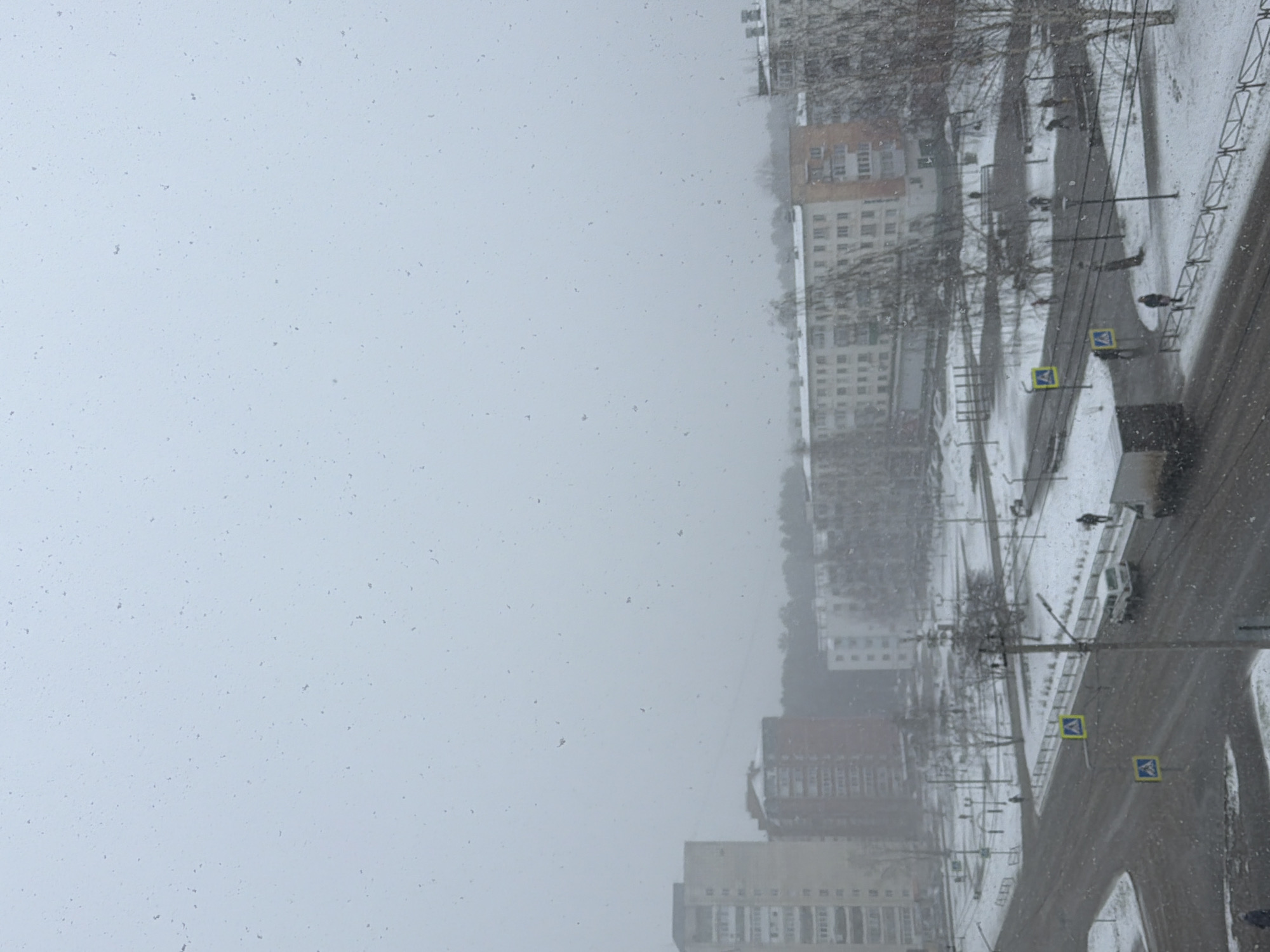 The image shows an overcast scene during a snowfall in Chaikovsky, Russia. The perspective is from an elevated position, likely a window, overlooking a street scene.
Buildings are visible on the right side of the image; these are multi-story residential structures. The street below is partially covered in snow. There are pedestrian crosswalk signs visible along the street.
Several people are walking along the sidewalk. There are also parked cars and a utility truck on the street. The bare trees suggest it is late fall or winter. The sky is a uniform gray, indicating heavy cloud cover and falling snow.