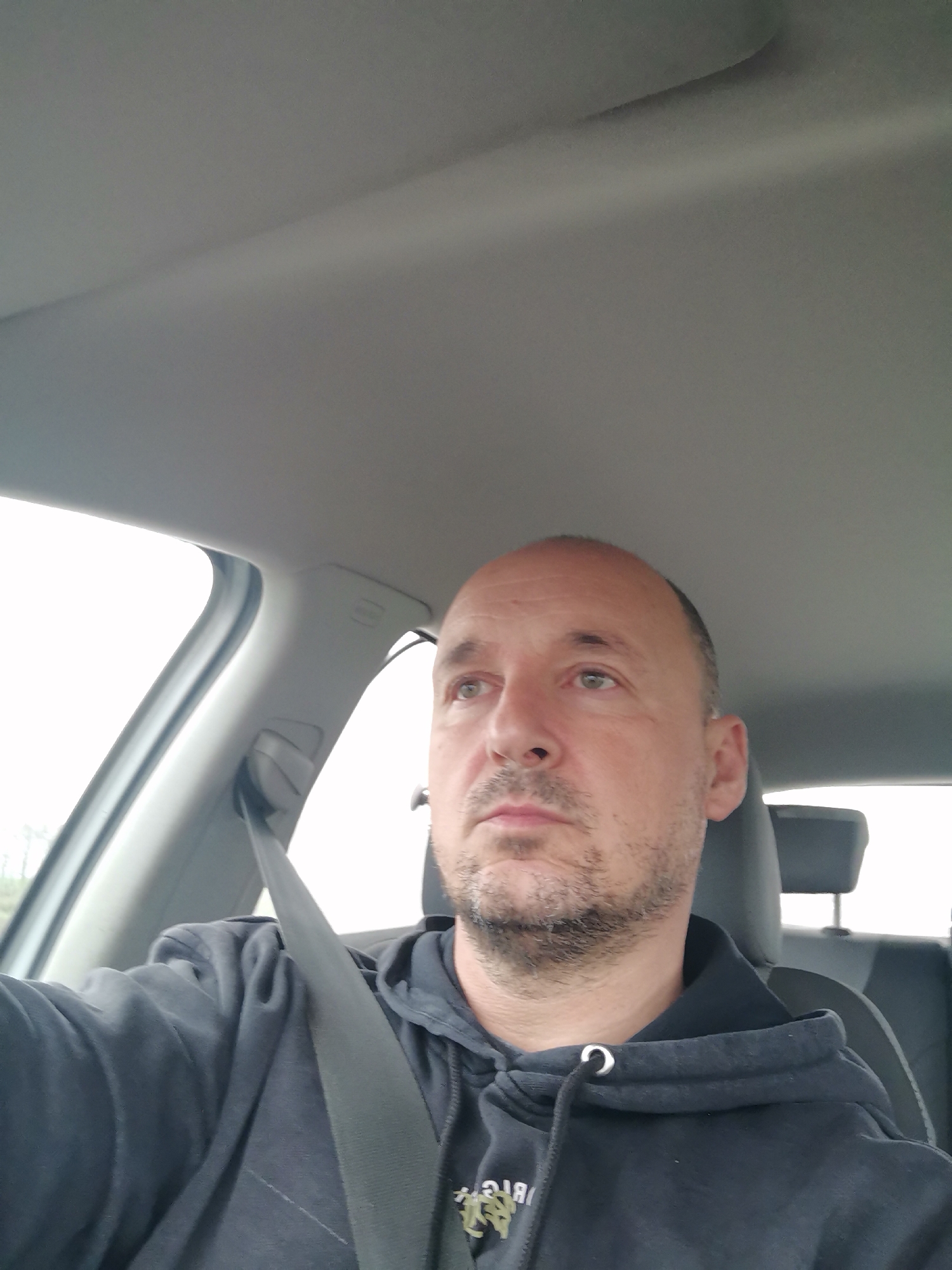 A man is depicted seated in what appears to be the driver's seat of a car. He is a middle-aged individual with a shaved head or very short hair on top, and short dark hair on the sides. He has a short beard or stubble and light-colored eyes. His expression is neutral, possibly pensive or focused, as he looks slightly upwards and to his right.
He is wearing a dark (likely black or dark blue) hooded sweatshirt with a visible drawstring at the collar. A black seatbelt crosses his chest, indicating he is secured in the vehicle. The interior of the car is visible, featuring a light grey headliner and A-pillar. The headrest of the seat is visible behind him.
Through the car's windows, a bright, light sky is visible, suggesting it is daytime and possibly overcast weather conditions. On the front of the man's hoodie, reversed text is visible, appearing as "ЯІЯ" (GIB reversed) and other indistinct characters, likely part of a logo or brand name.
The scene takes place inside a vehicle, possibly in motion or parked. The given location context for the image is Branishte, Bulgaria.