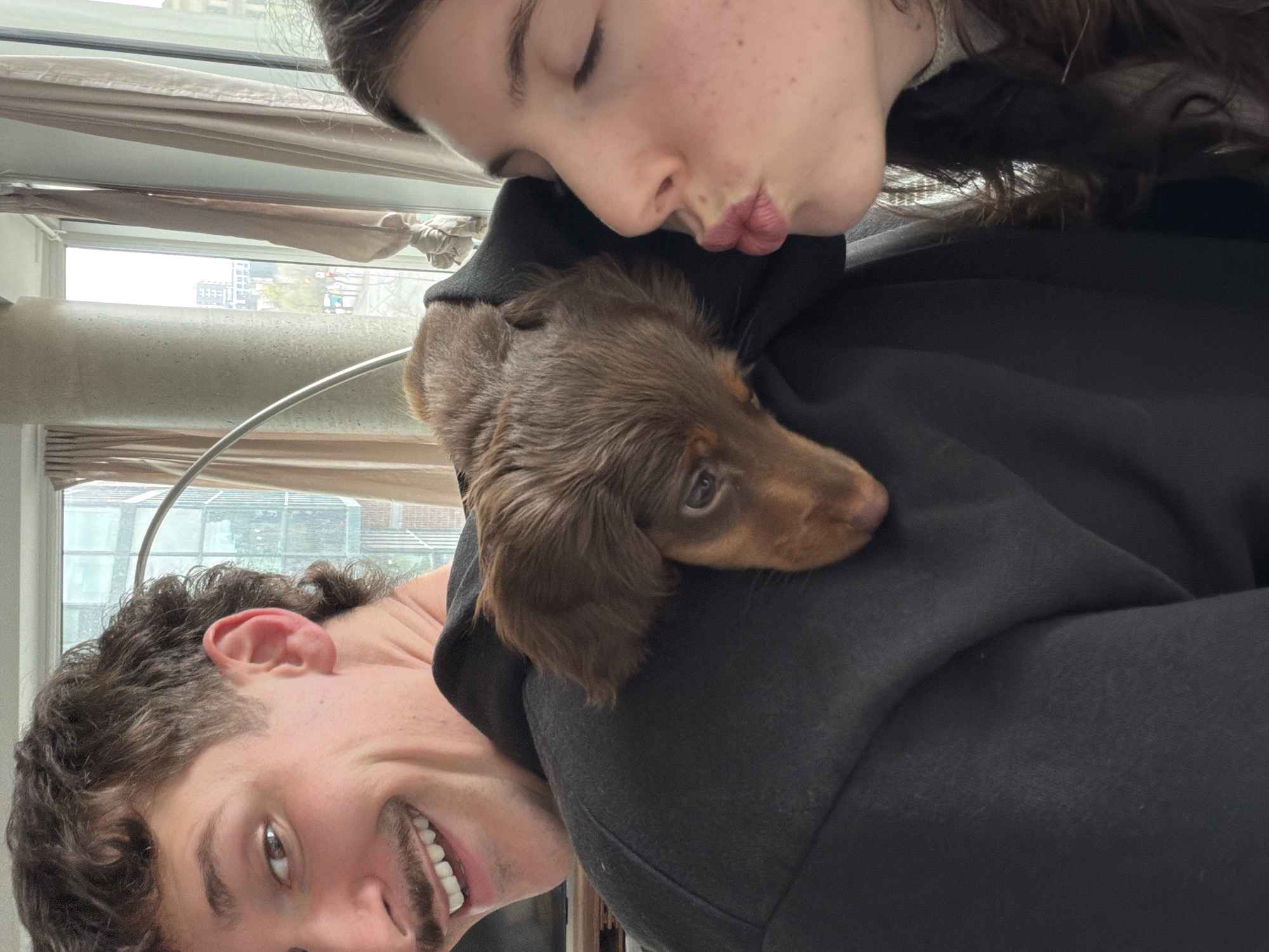 The image shows a selfie taken indoors, with the scene oriented sideways. A young man with brown curly hair and a mustache is smiling at the camera from the lower-left corner. A young woman is leaning over him, her eyes closed and lips pursed as if to kiss a small, brown, long-haired dachshund puppy resting on the shoulder of a black hoodie. A window is visible in the background, revealing a cityscape likely in Montreal, Canada, identifiable by the architectural style of the buildings visible through the window. The lighting suggests it is either daytime or that the photo was taken in a well-lit room. The focus is on the people and the dog, emphasizing an affectionate interaction.