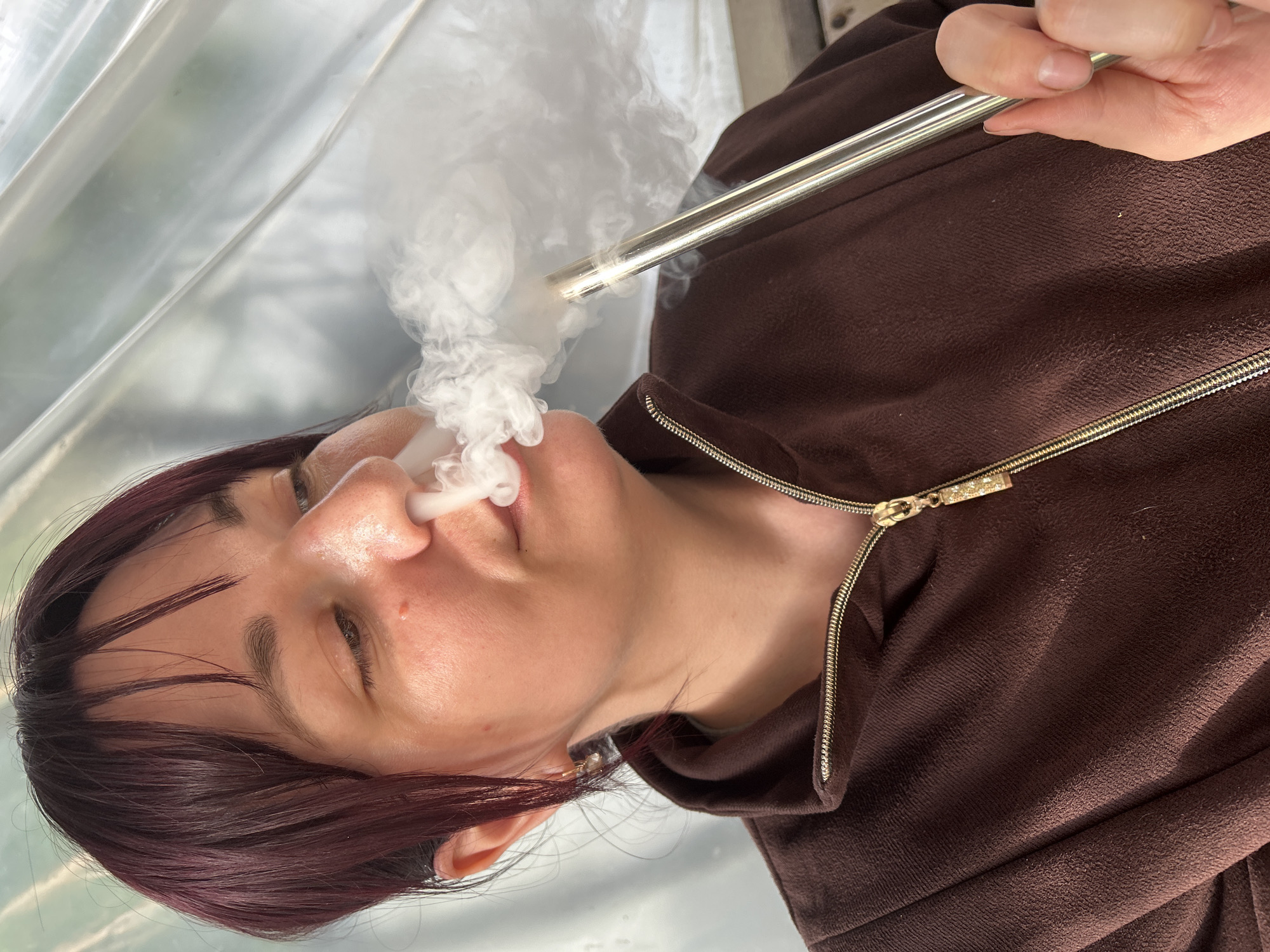 In Fokino, Russia, a woman is depicted from the chest up, engaged in exhaling or inhaling a significant cloud of white vapor from a metallic device. She has dark reddish-brown hair styled with bangs and fair skin, showing a small reddish blemish on her upper right cheek. Her eyes are partially closed and directed downwards. She wears a brown, textured, possibly fleece or corduroy-like jacket with a gold-colored zipper and a decorative, rectangular gold zipper pull. A thin, reflective, silver-toned metallic tube, resembling a hookah hose mouthpiece or a large vape pen, is held to her mouth, with her right hand partially visible gripping the tube. A substantial plume of white vapor emanates from her mouth and partially from her nose, spreading across the right side of the frame. A small gold-colored hoop earring is visible in her left ear. The background consists of a light-colored, possibly translucent, and slightly wrinkled or corrugated material, exhibiting diagonal folds, suggesting an interior or sheltered outdoor setting.