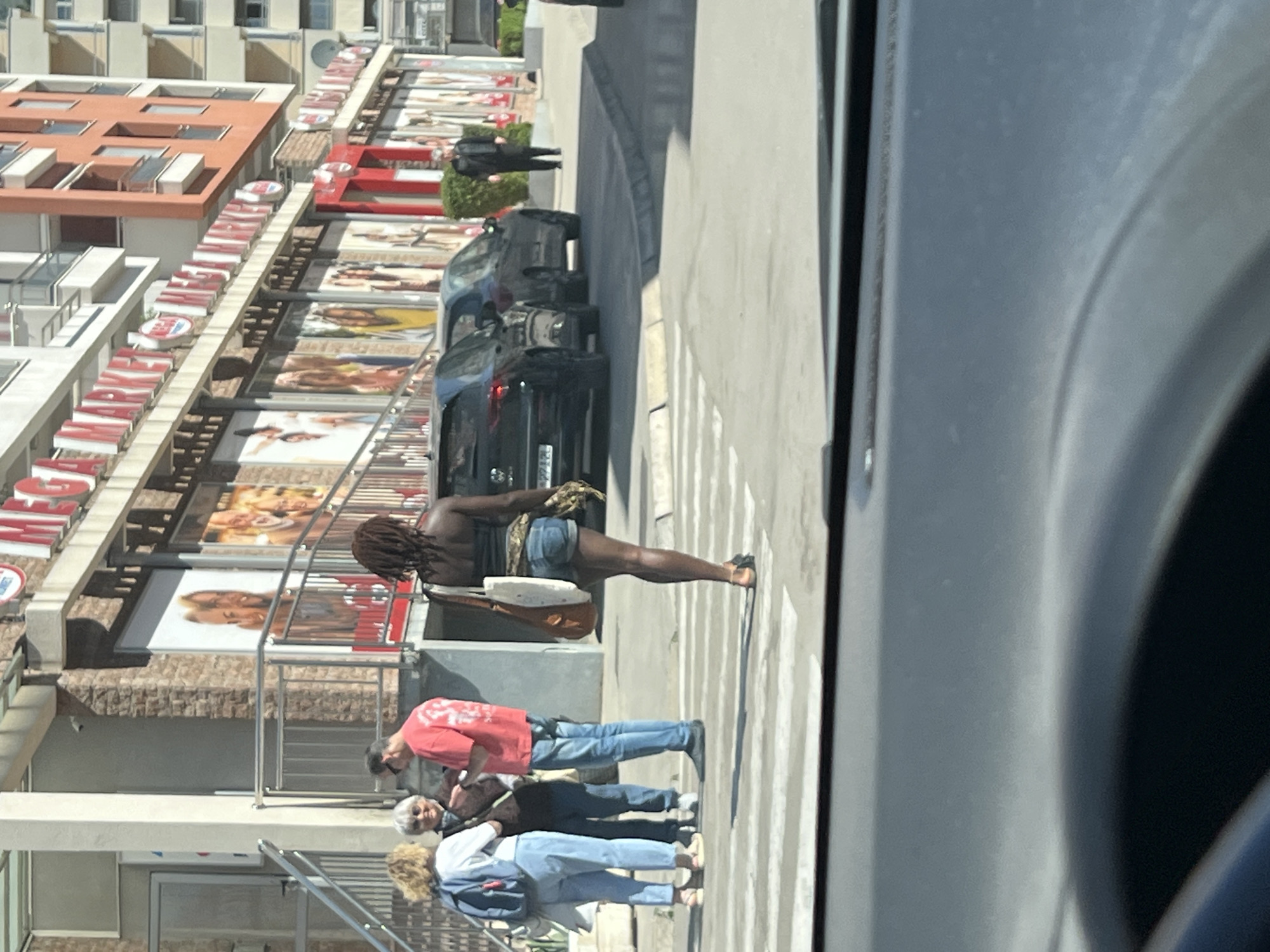 This image captures a vibrant street scene in Budva, Montenegro, under bright daylight. The photo is taken from inside a vehicle, looking out onto a street and sidewalk lined with buildings.
In the immediate foreground, center-right, a person with dreadlocks, wearing a dark top (possibly shirtless) and denim shorts, stands on a crosswalk. They are carrying a light-colored, possibly white, shoulder bag with a distinct shape that suggests it might hold a musical instrument like a guitar.
Further left, on the sidewalk closer to the buildings, a group of three other pedestrians is visible. One person wears a red t-shirt and blue jeans. Next to them, an older woman with white hair and sunglasses walks alongside another person with blonde/light brown curly hair, wearing light blue jeans and carrying a backpack.
The buildings lining the street are multi-story, featuring balconies and prominent red-tiled roofs, characteristic of coastal Mediterranean architecture. The ground floors appear to house commercial establishments, indicated by large windows and numerous advertisements. Along one building, a long, illuminated sign with red letters spells out "MEGA", followed by more letters, suggesting a store like "MEGAMARKET". Below this, several large billboards display images of people, likely models or product advertisements.
On the street, a black SUV or car is parked, and part of its license plate, "4221", is visible. Another dark car is parked behind it. Strong shadows suggest it is midday or early afternoon on a sunny day.