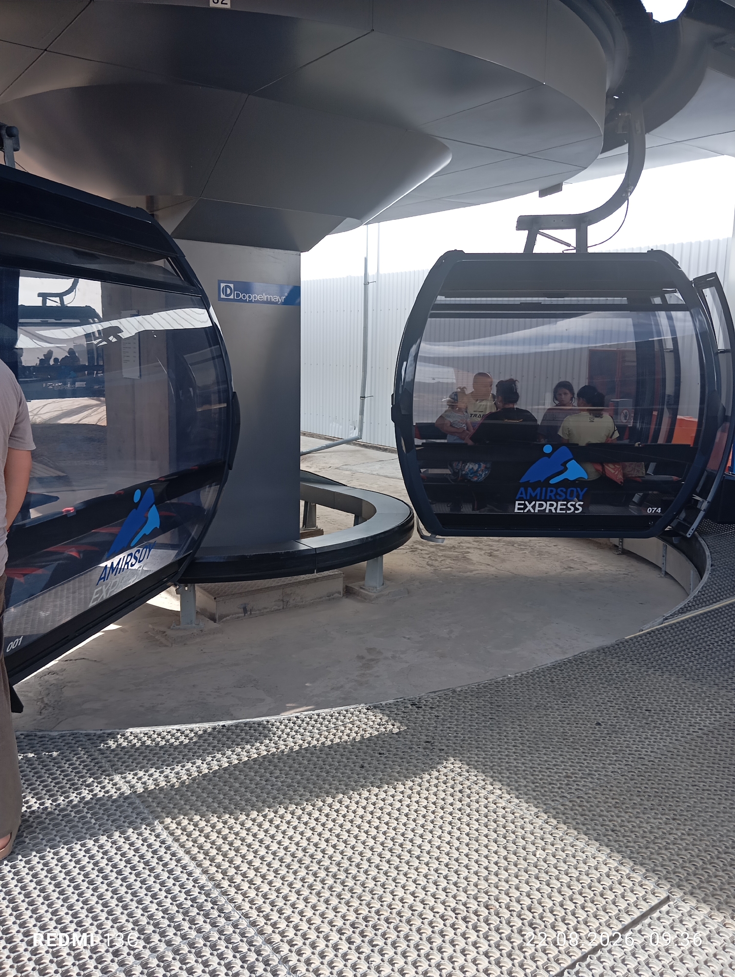 The image shows a gondola of the Amirsay Express cable car in Amirsay, Uzbekistan. The gondola is black with a large panoramic window and seats inside. It is stopped at a station, with people visible inside the gondola. The station structure is dark grey and modern, with a Dopplemyr logo visible on a pillar. The ground is paved with a textured mat. The overall scene suggests a sunny day at a ski resort or mountain attraction.