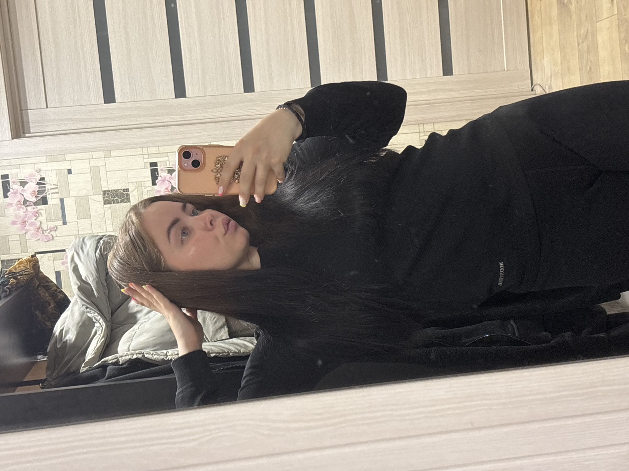 A young woman is taking a selfie in front of a mirror. She is lying down, with her head positioned towards the left side of the frame. She has long brown hair, fair skin, and is wearing a black outfit. Her phone, which has a decorative case, is held up in her right hand. The background shows a room with a light wood-paneled wall and a decorative tiled section with a floral pattern. There is also a gray jacket visible behind her head. The photograph was taken in Izhevsk, Russia.