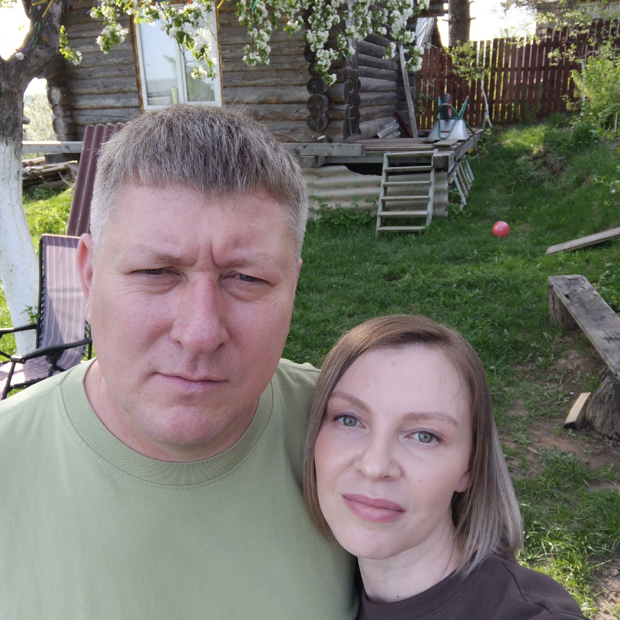 The image shows a man and woman posing for a selfie outdoors. The man is on the left wearing a green t-shirt, with short gray hair and a serious expression. The woman is on the right, wearing a dark brown t-shirt, with shoulder-length light brown hair.
Behind them is a wooden cabin with a window and flowering branches. A set of steps lead to a small porch. In the background, there is a wooden fence, green grass, and some other objects including a red ball. The presence of a typical Russian cabin style house and outdoor landscape suggests the location may well be in Chaikovsky, Russia.