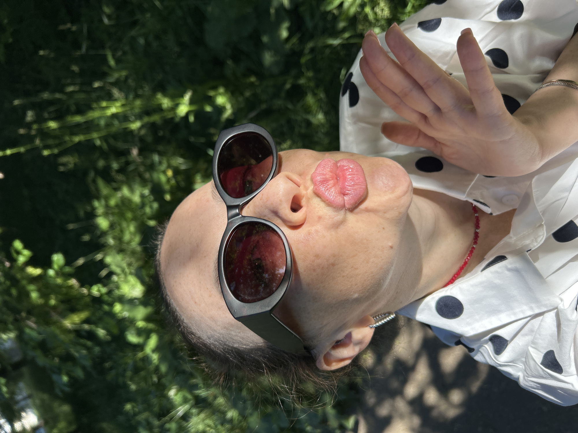 The image is a close-up of a woman outdoors in what appears to be a park or natural area, with lush green foliage in the background. The woman is wearing large, dark sunglasses and a white shirt with black polka dots. She has her lips puckered as if to blow a kiss, and her right hand is raised with fingers spread. She is also wearing a red beaded necklace and a silver earring. The lighting suggests it is a sunny day, casting shadows on the ground and the woman's face. The location is identified as Chaikovsky, Russia.