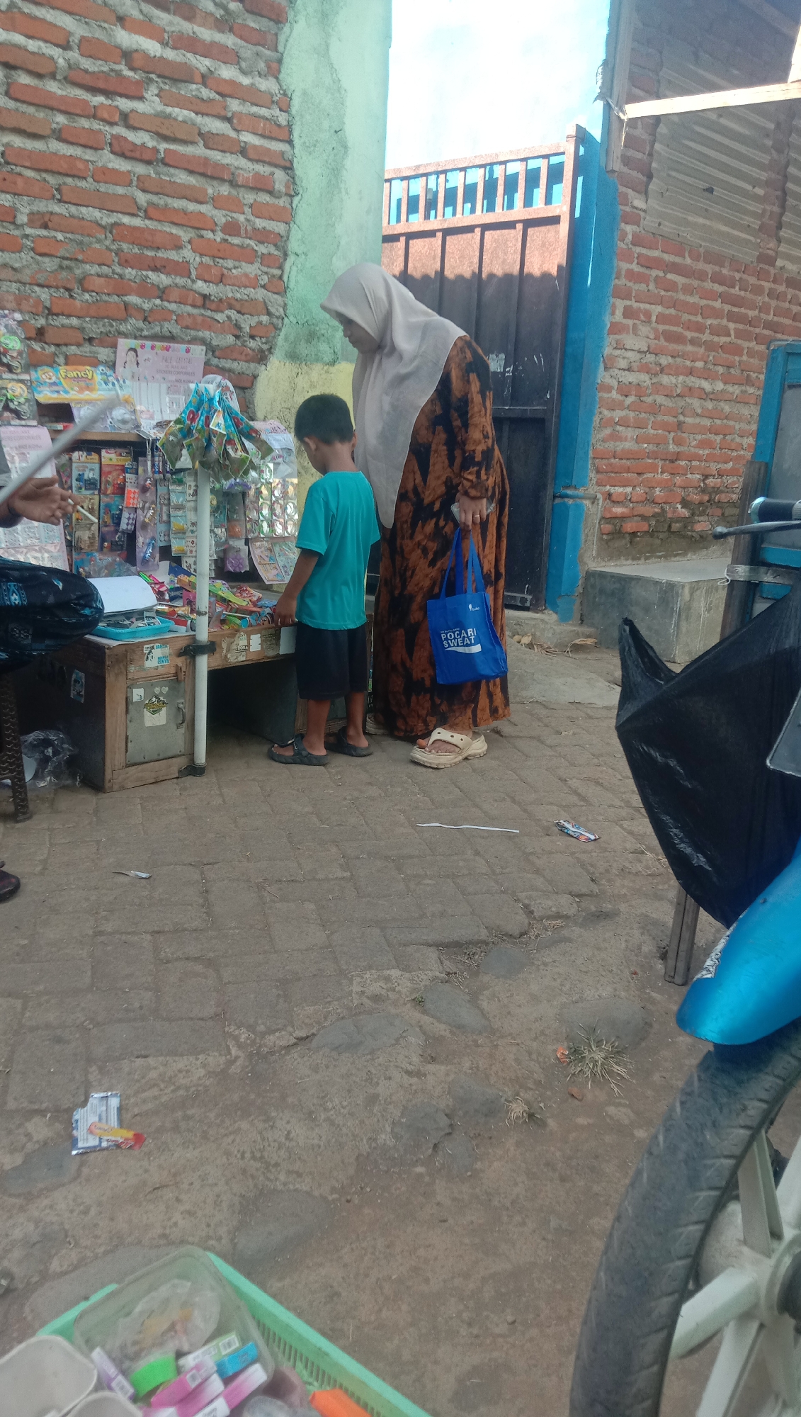 A woman and a boy are standing at a makeshift stall displaying various items, possibly snacks or toys. The woman is wearing a beige hijab and a patterned dress, and is holding a blue shopping bag with the words "POCARI SWEAT" printed on it. The boy is wearing a blue t-shirt and black shorts. They appear to be interacting with the stall's merchandise. The stall is set up on a paved area, with a brick wall in the background. The scene suggests a street-side market or shop in Karang Anyar, Indonesia. The environment appears to be somewhat cluttered, with debris on the ground. A portion of a motorcycle is visible in the foreground.