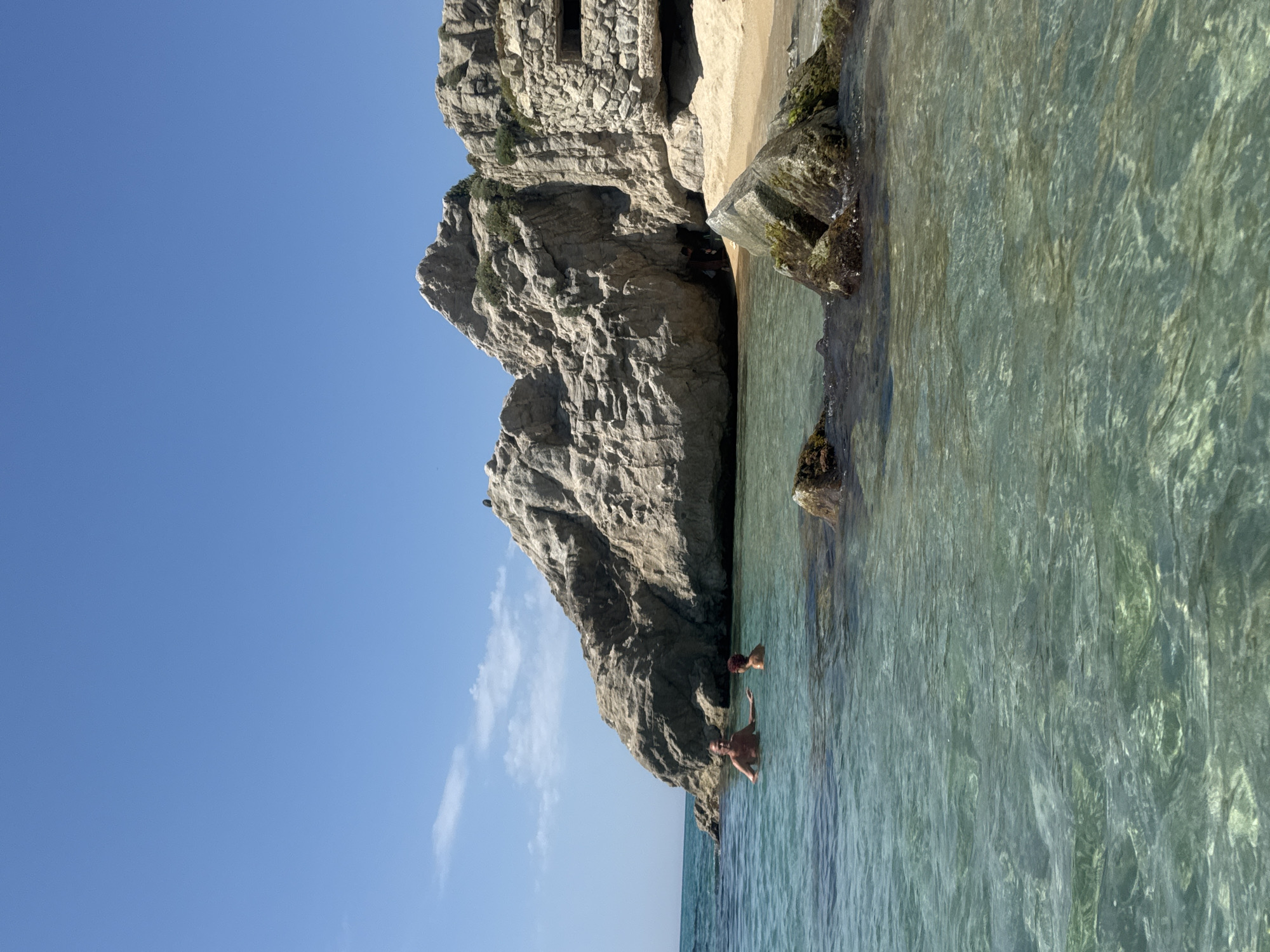 The image depicts a scene at the "Búnkers del Turó de la Bota" in Arenys de Mar, Spain. Two individuals are partially submerged in the clear turquoise water, facing towards the camera. The man on the left is older, with grey hair and a tanned complexion, and is looking towards the woman on his right. The woman has reddish-brown hair and is also in the water. Behind them, a large rocky formation dominates the landscape, with a sandy beach visible at its base. Part of a concrete structure, likely a bunker, is integrated into the rock face on the right side of the image. The sky is a clear, bright blue with a few wispy clouds. The water is shallow and transparent, allowing the seabed to be visible in places. Rocks are scattered in the foreground of the water.