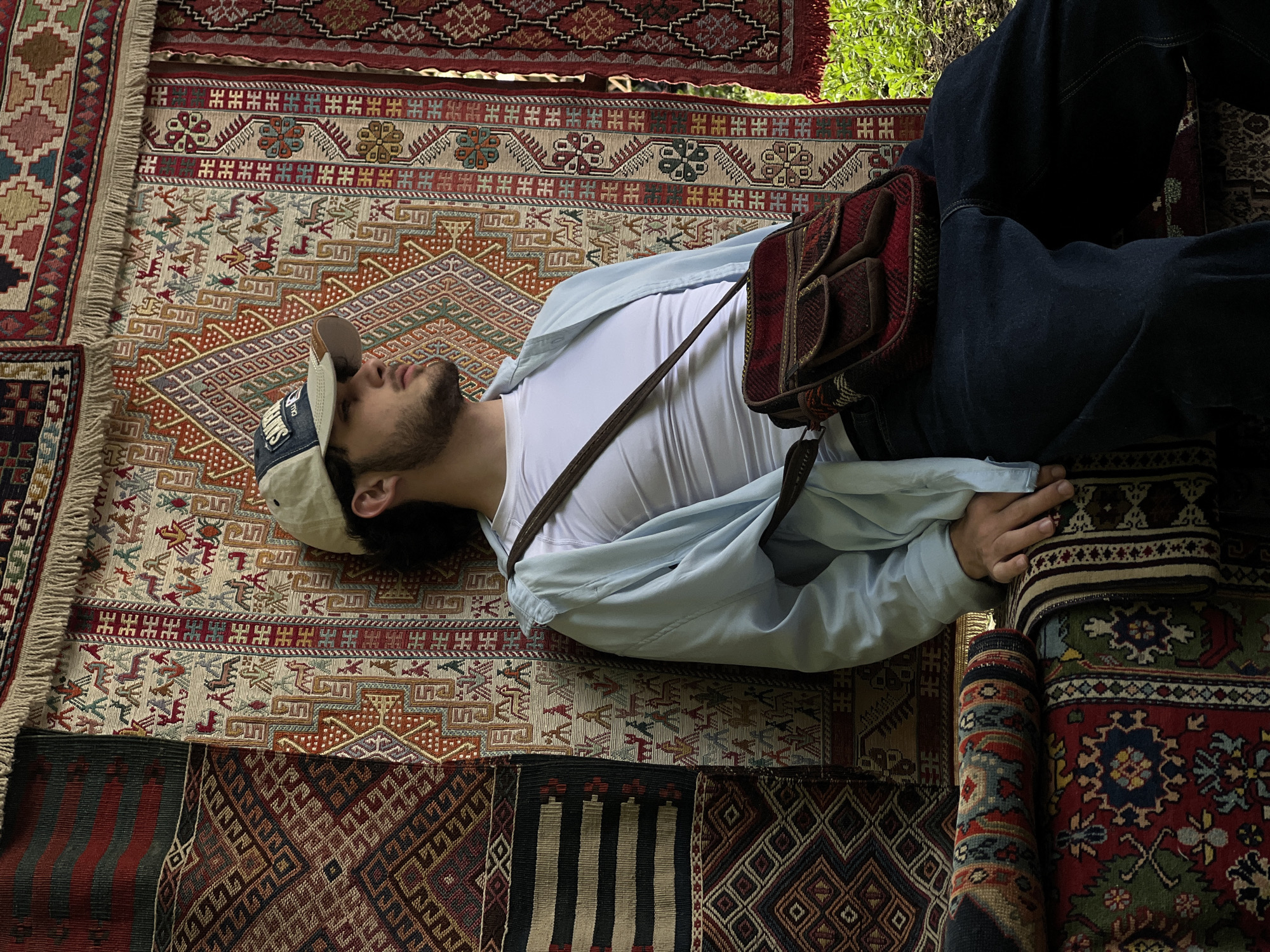 The image shows a person lying on a collection of intricately patterned rugs. The individual, wearing a white t-shirt, a light blue shirt, dark jeans, and a baseball cap, is positioned diagonally across the frame, with their head near the upper left corner and legs extending towards the lower right. A patterned bag rests on their torso. The rugs beneath the person are diverse in design and color, featuring geometric motifs, floral patterns, and fringed edges. The top of the image shows trees and bright green leaves. The city of Yerevan, Armenia is not discernible from the image.