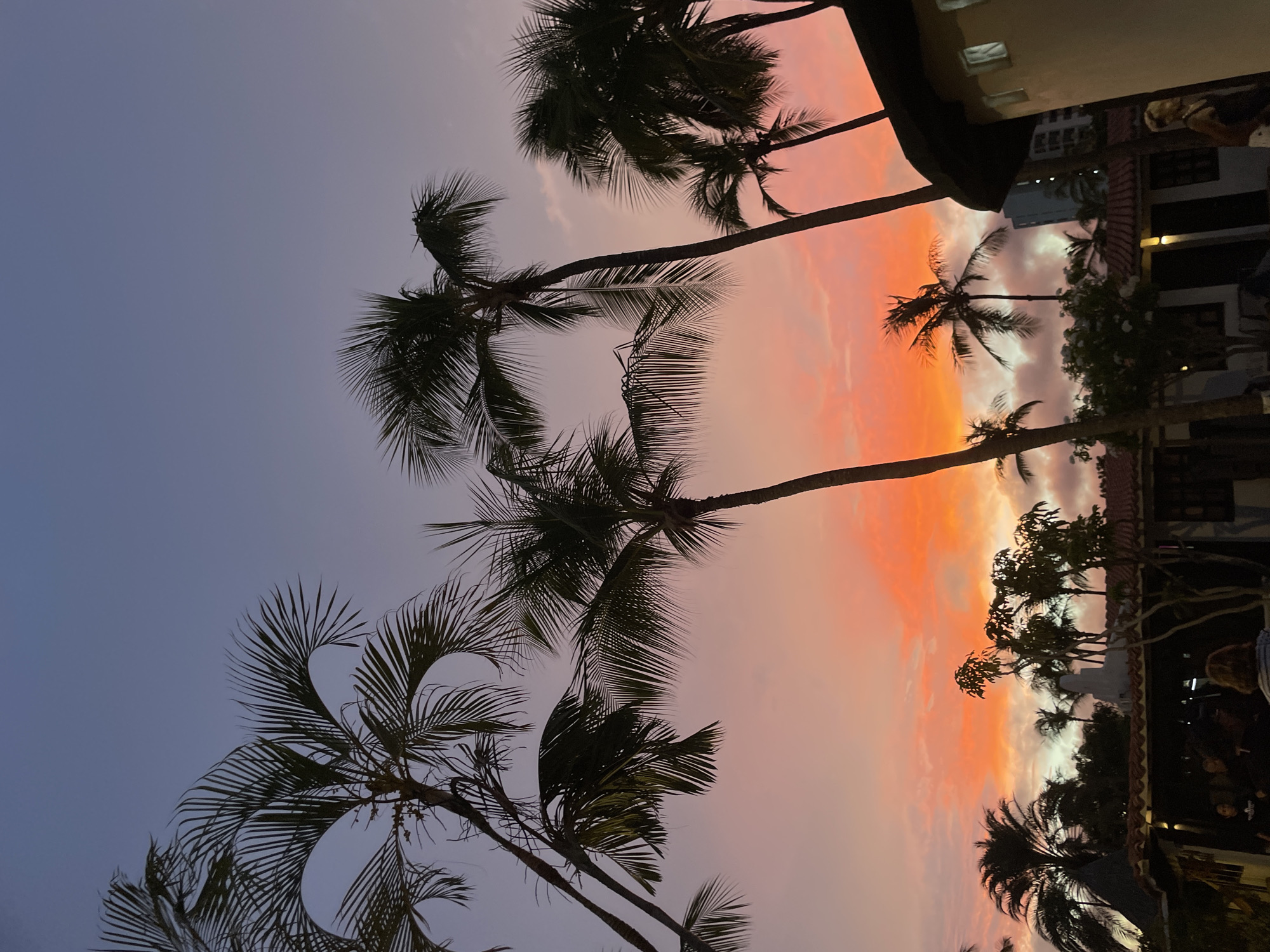 The image depicts a vibrant sunset scene with silhouettes of palm trees against a gradient sky of soft purples and fiery oranges. In the foreground, a building with a tiled roof and warmly lit windows is visible, suggesting a resort or restaurant setting. Several people are gathered under the covered patio area of the building, their forms indistinct in the low light. Further in the background, more palm trees and a modern high-rise building can be observed, indicating an urban coastal environment. This scene is likely located in Porlamar, Venezuela, a city known for its beautiful beaches and tropical atmosphere, where such colorful sunsets are a common spectacle.