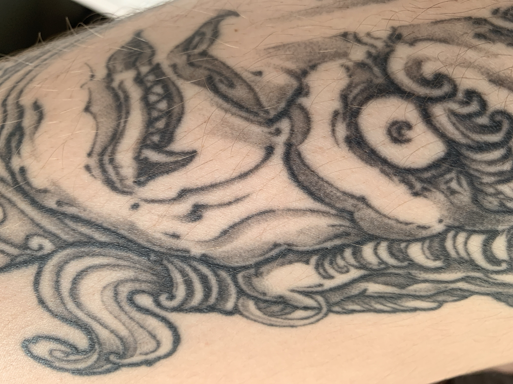 The image displays a close-up of a tattoo on human skin. The tattoo is rendered in black ink and depicts a stylized face with swirling hair or a headdress. The details of the tattoo include sharp teeth, a prominent brow, and decorative curls. The skin is light-toned and appears to have fine hairs. The image is framed from a slightly angled perspective, focusing on the texture of the skin and the linework of the tattoo. The background is indistinct and out of focus. There is no information in the image to suggest a location in Stavropol, Russia.